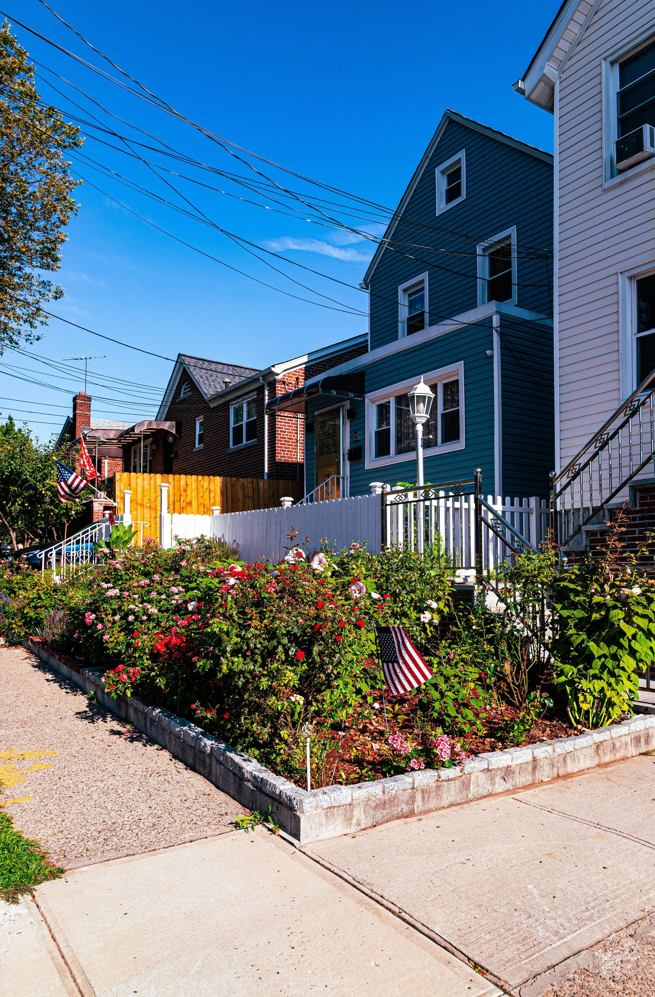 Colorful front yards of houses with American flags, blooming flowers, and a bright blue sky.