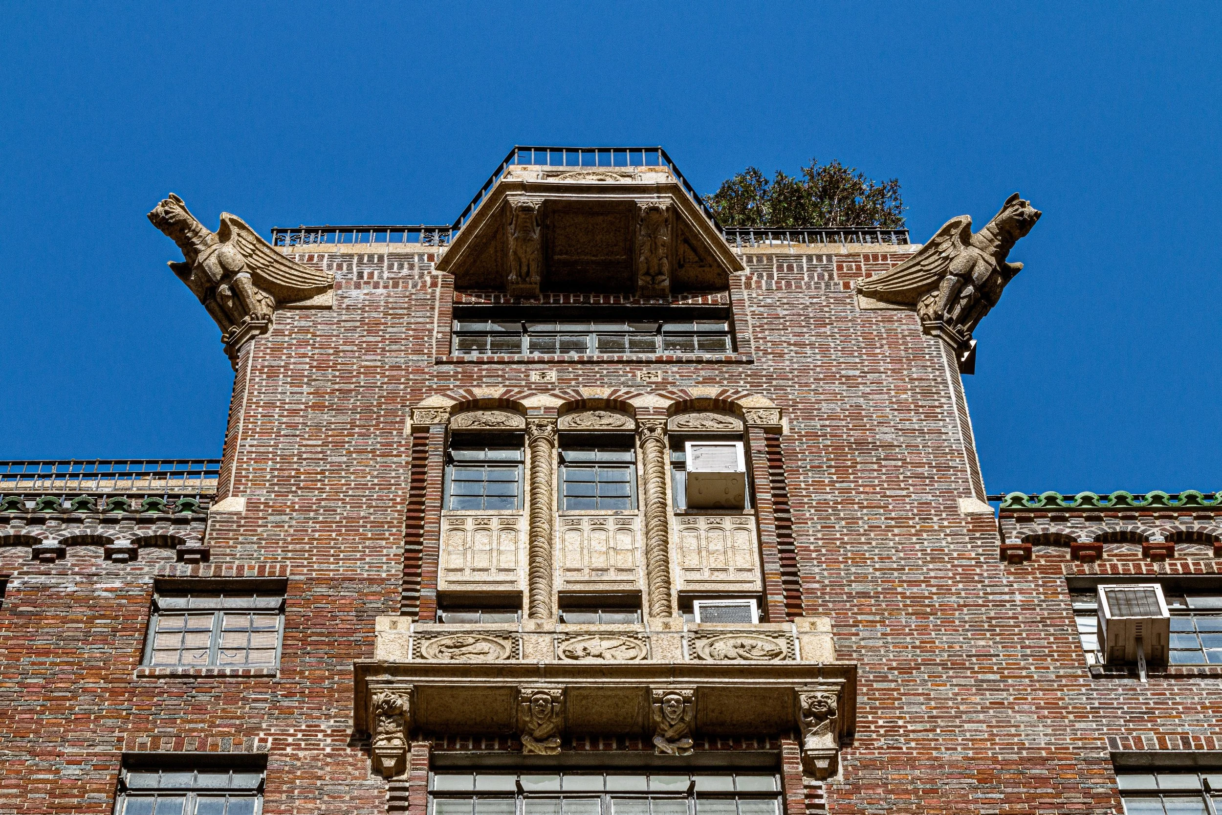 Close-up of a historic brick building with ornate stone architectural details, including two gargoyle sculptures on each side at the top, and a decorative balcony with sculpted figures and carvings beneath the windows, set against a clear blue sky.