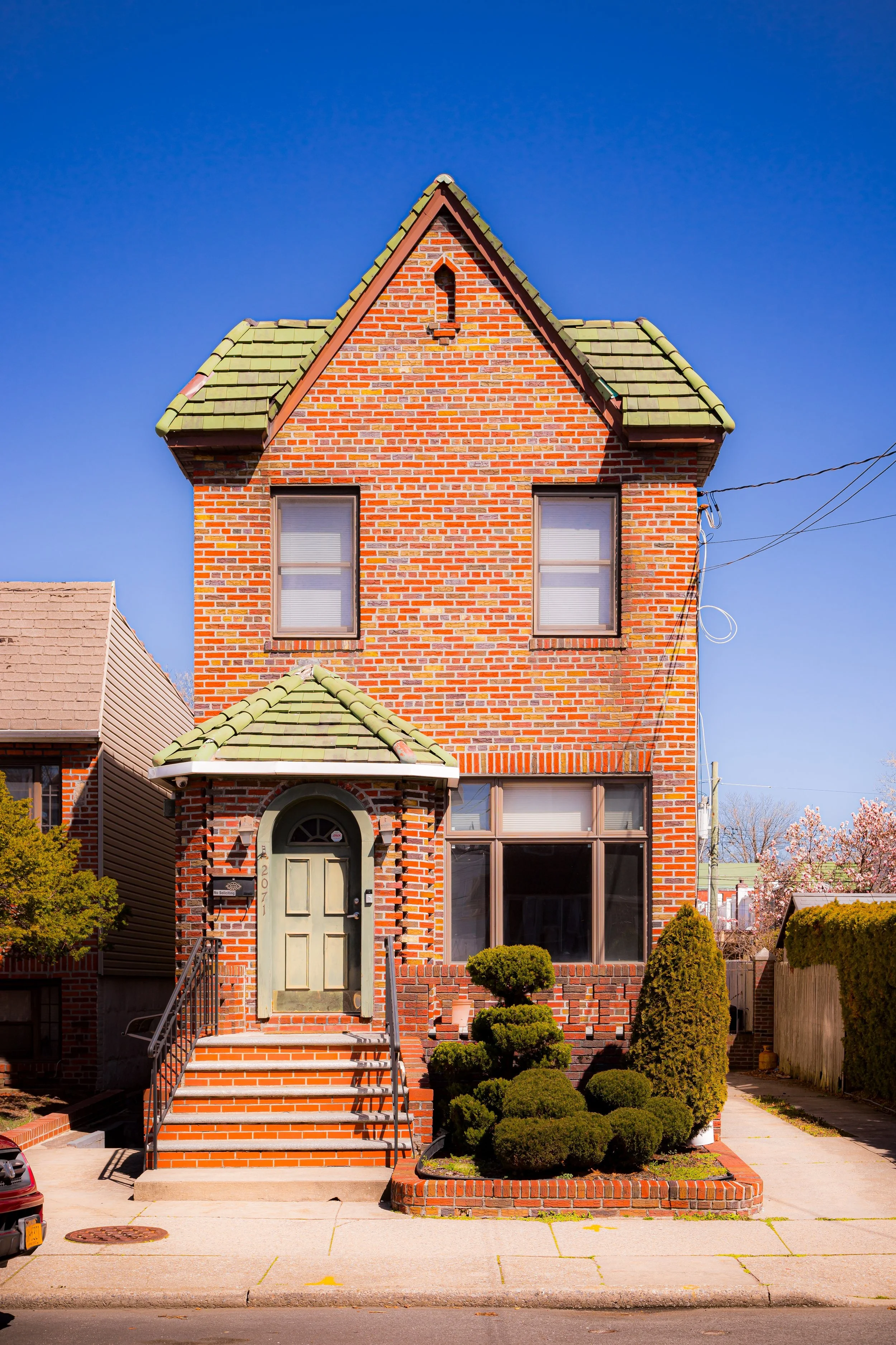A tall, red brick house with a green tiled roof, a small arched front porch, steps, and neatly trimmed bushes in the yard.