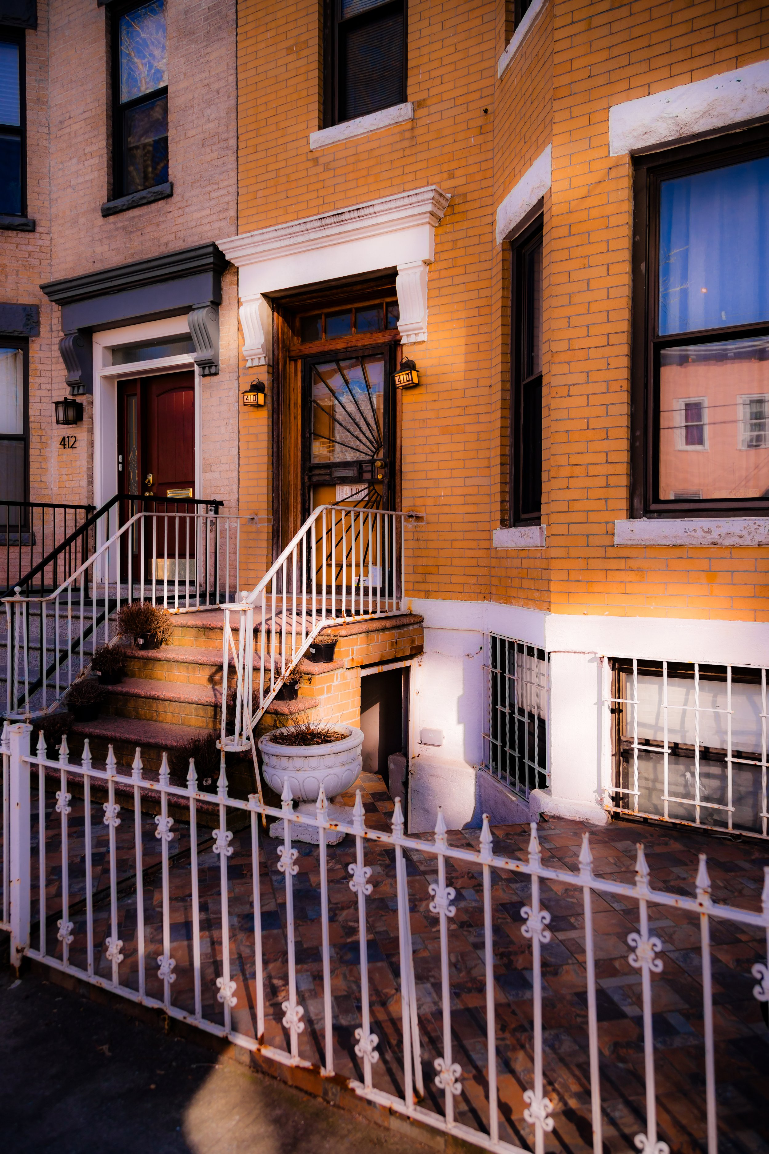 Close-up of the front entrance of a brick townhouse, featuring a dark wooden door with metal detail, white decorative crown molding, a white front railing, and small potted plants on the steps, with street and neighboring building reflections visible in the windows.