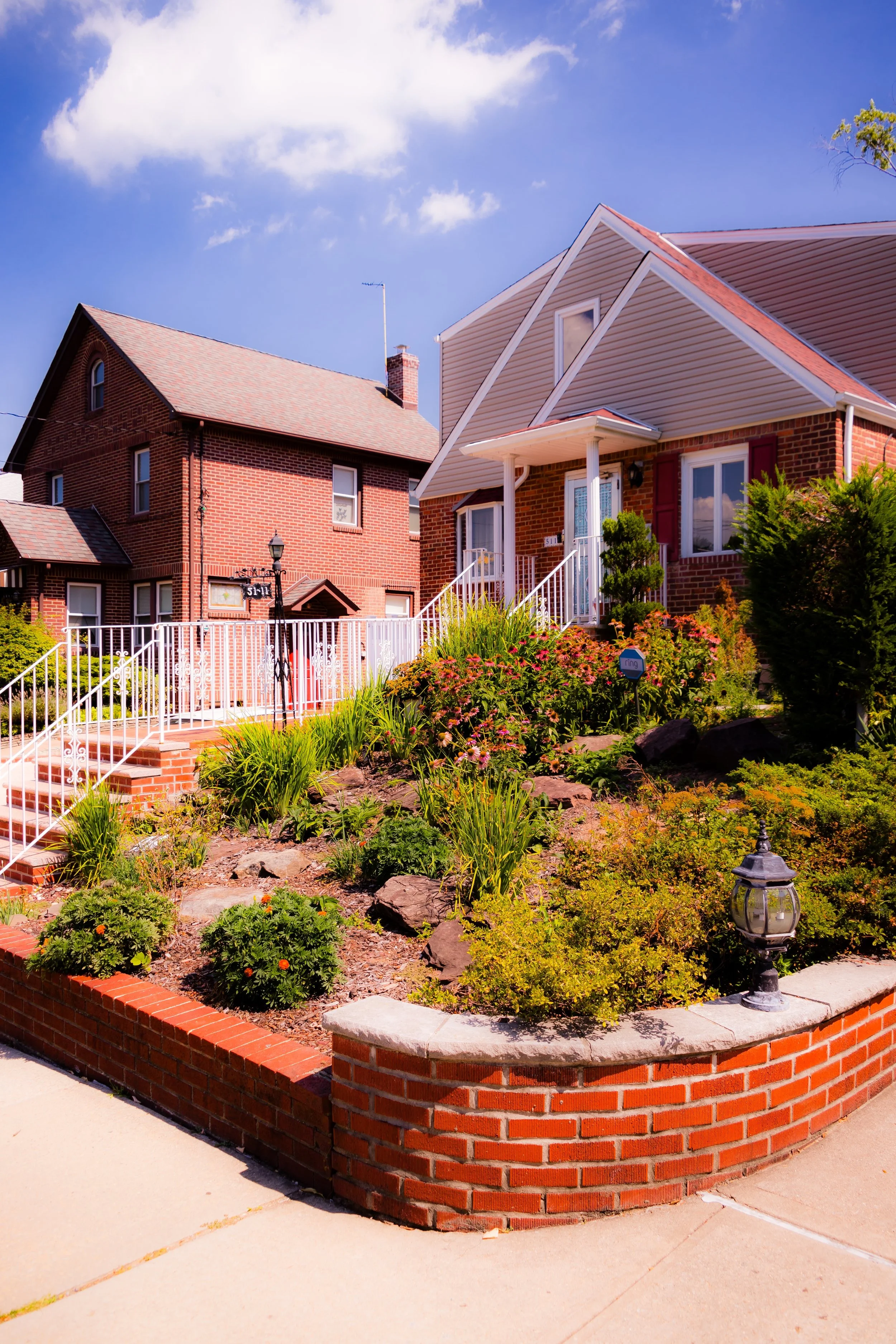 A residential front yard with a garden, brick wall, and sidewalk. Two houses are visible, one brick and one with siding, under a blue sky with some clouds.