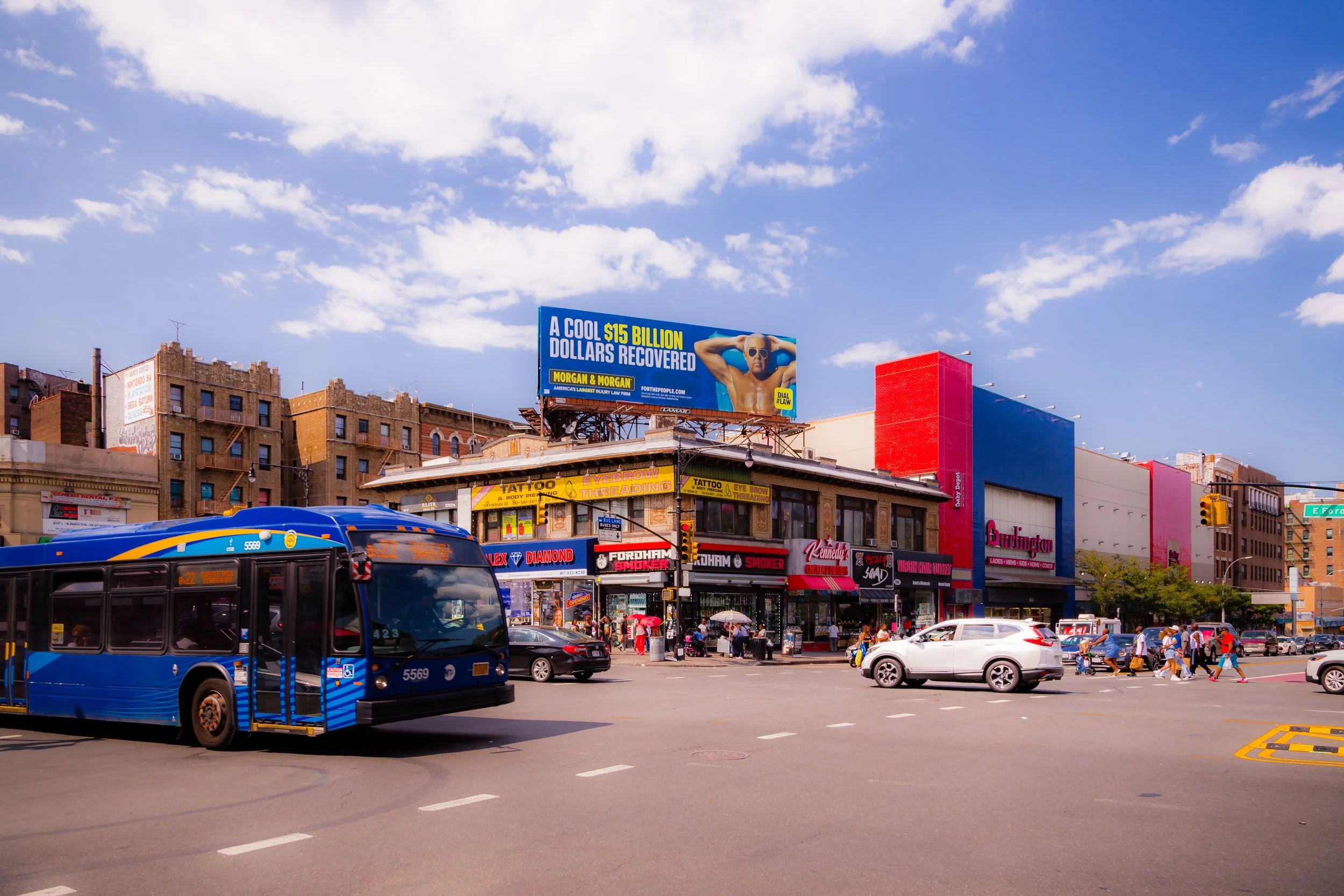 Urban street scene with a blue city bus, cars, and pedestrians crossing the intersection under a bright blue sky with scattered clouds, storefronts, a billboard, and a traffic light.