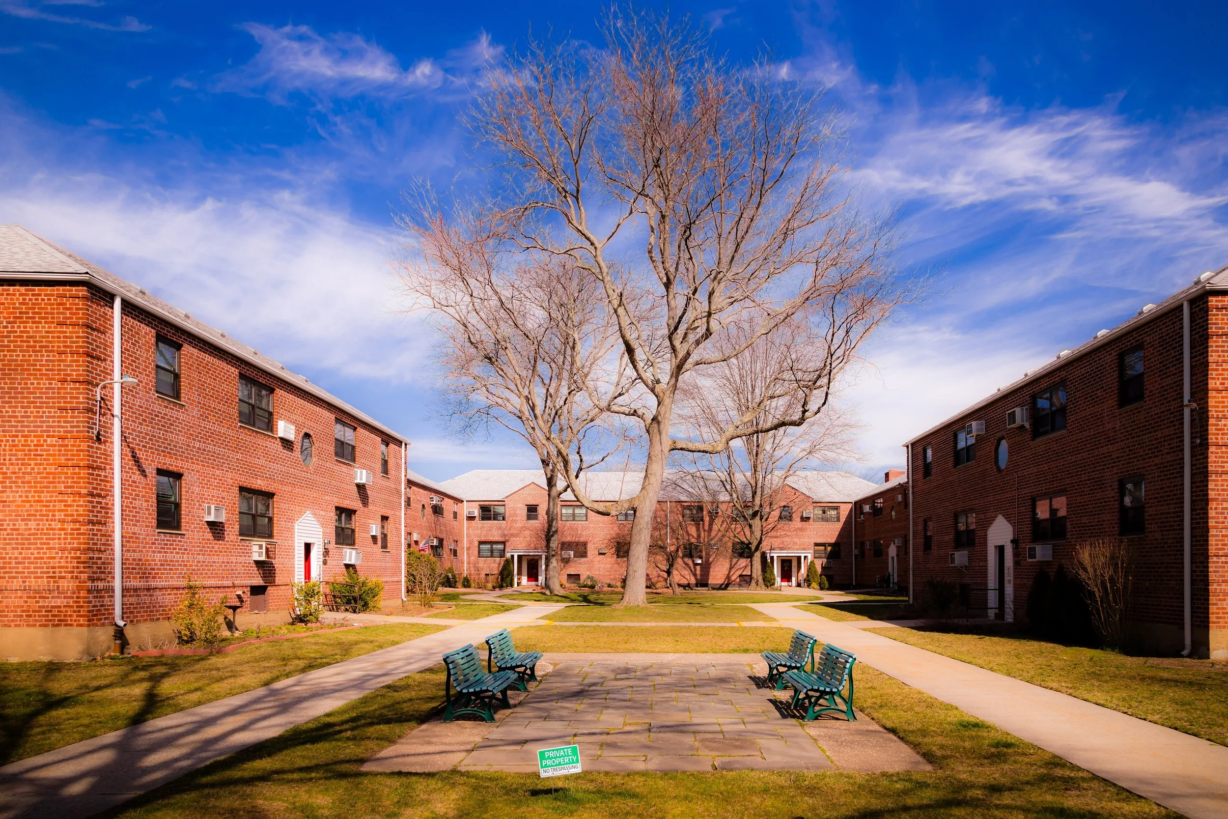 Courtyard with leafless trees, brick apartment buildings, and green benches under a blue sky with white clouds.