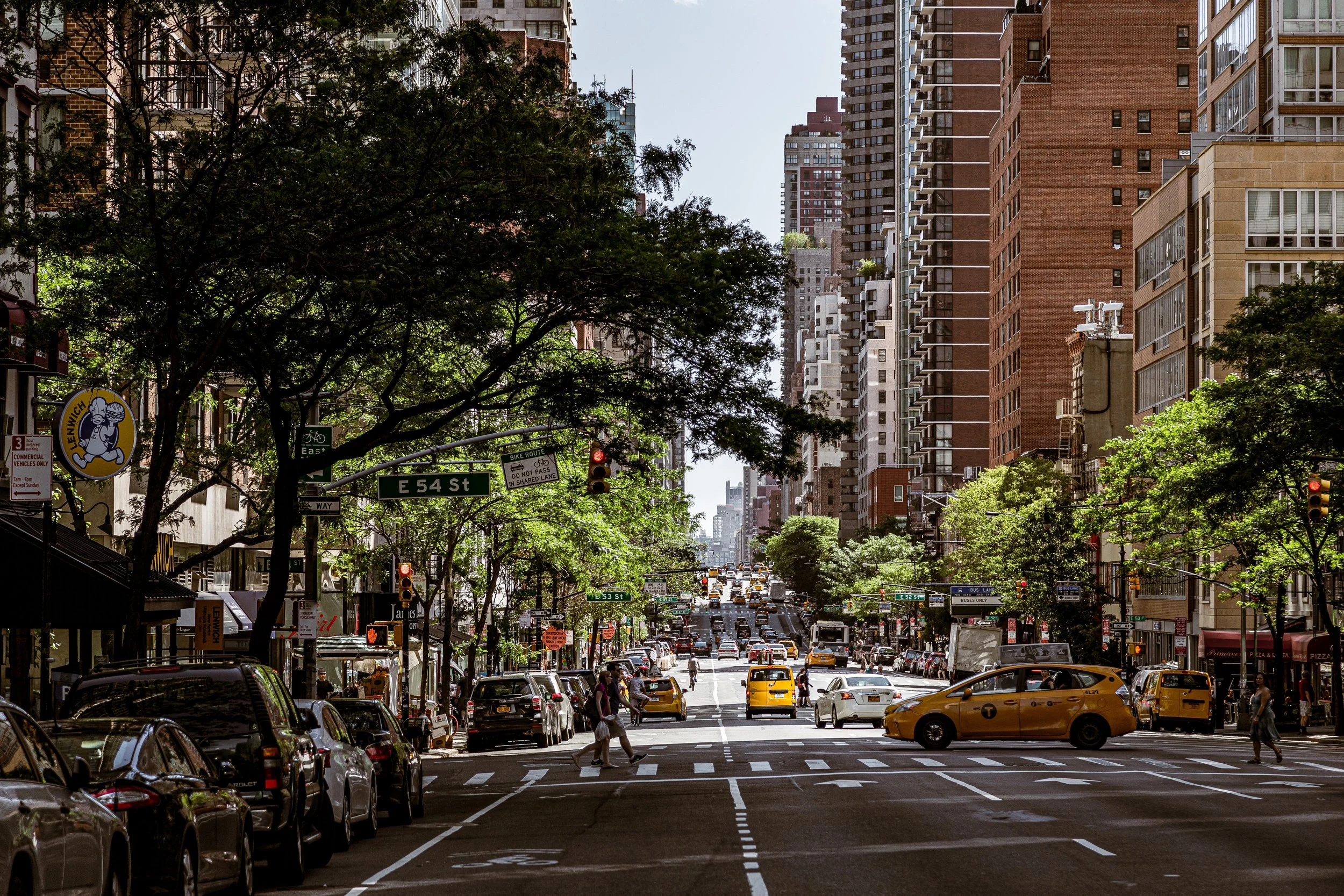 City street scene with tall buildings, trees, cars, taxis, and pedestrians at an intersection, with signage including E 54 St and nearby shops.
