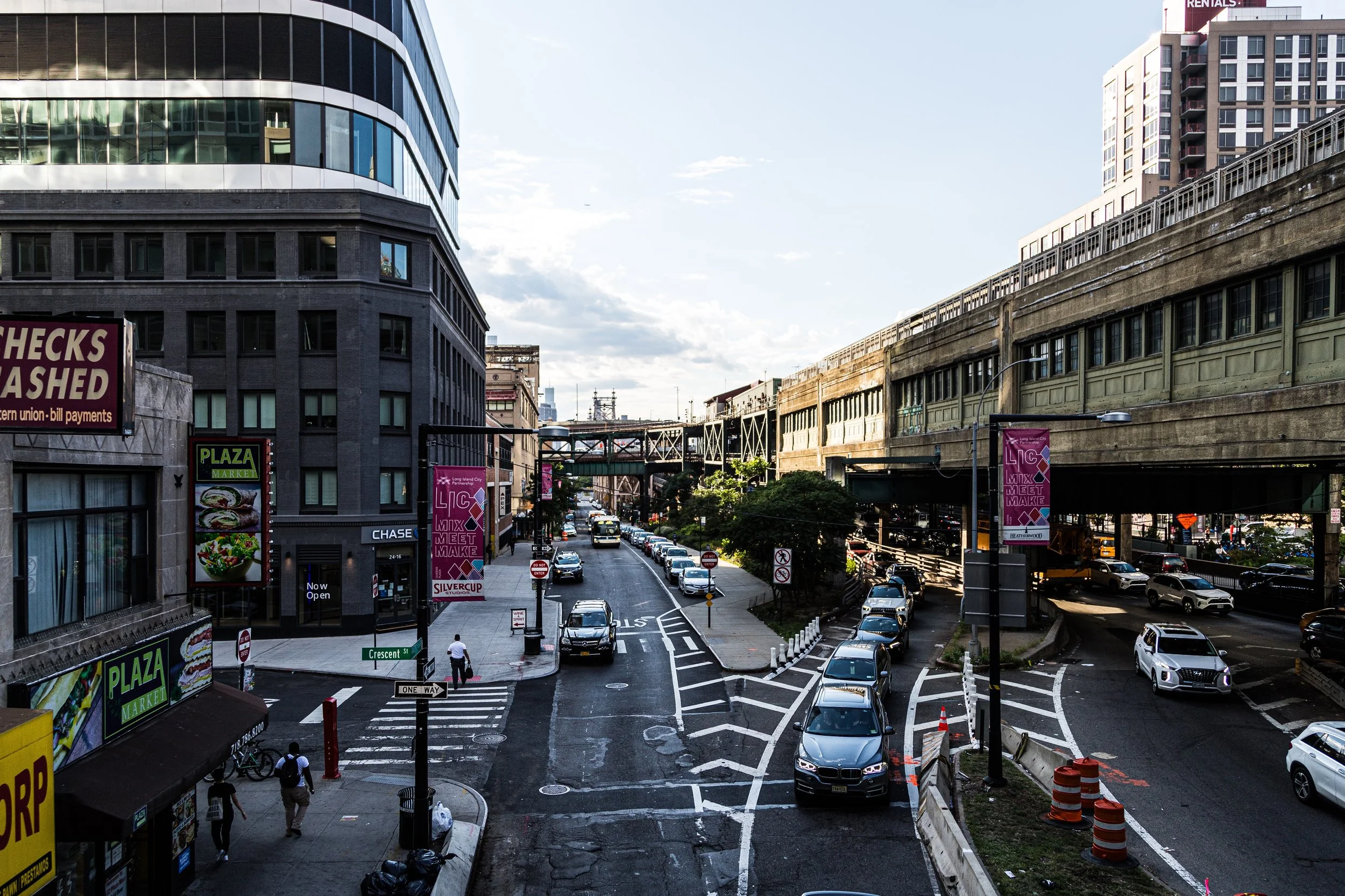 Urban street scene with cars parked along the curb, pedestrians walking, a multi-story building with glass windows on the left, elevated train tracks on the right, and various signs and advertisements visible