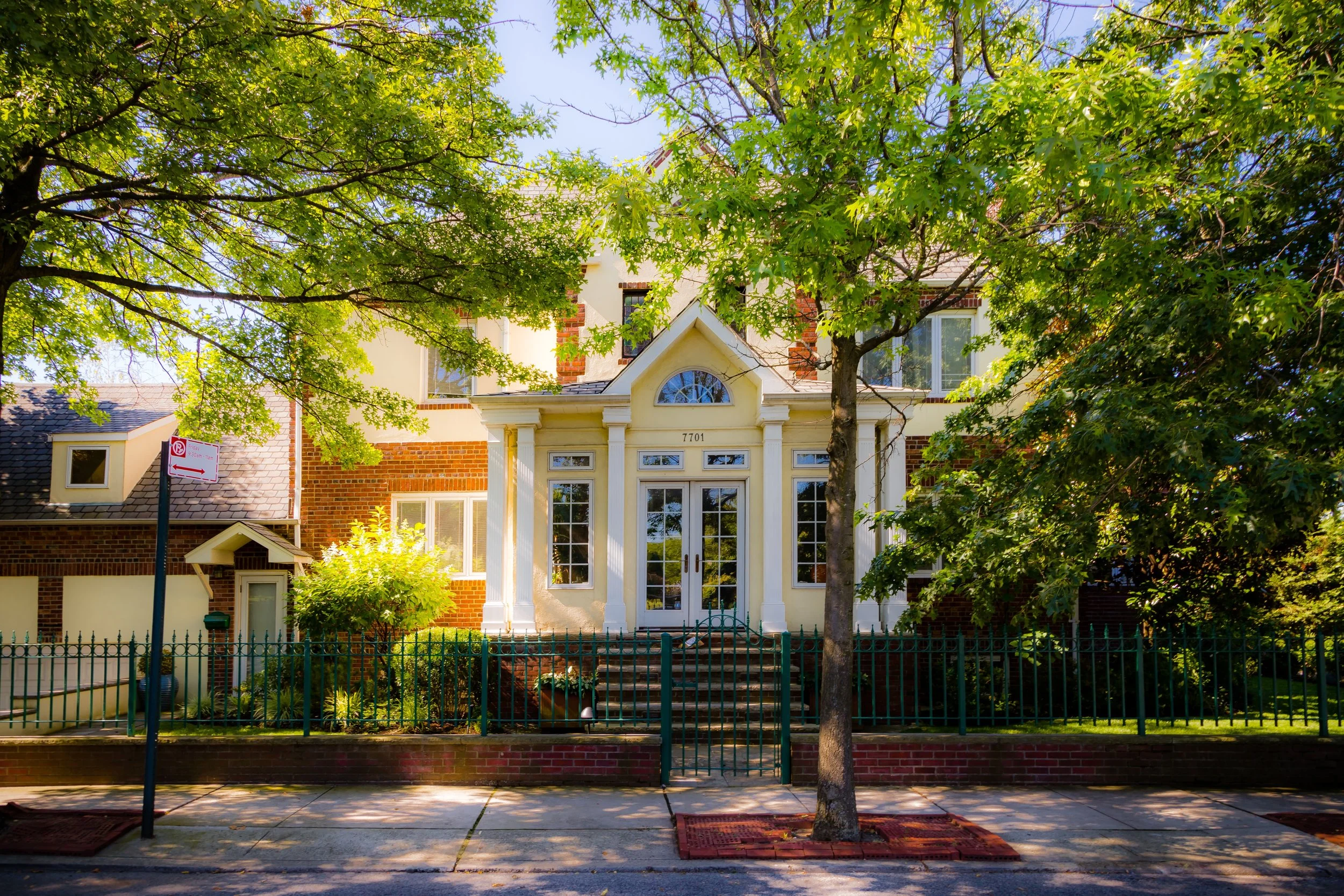 A house with a white facade and red brick accents, surrounded by green trees and a green fence, with a sidewalk in front.