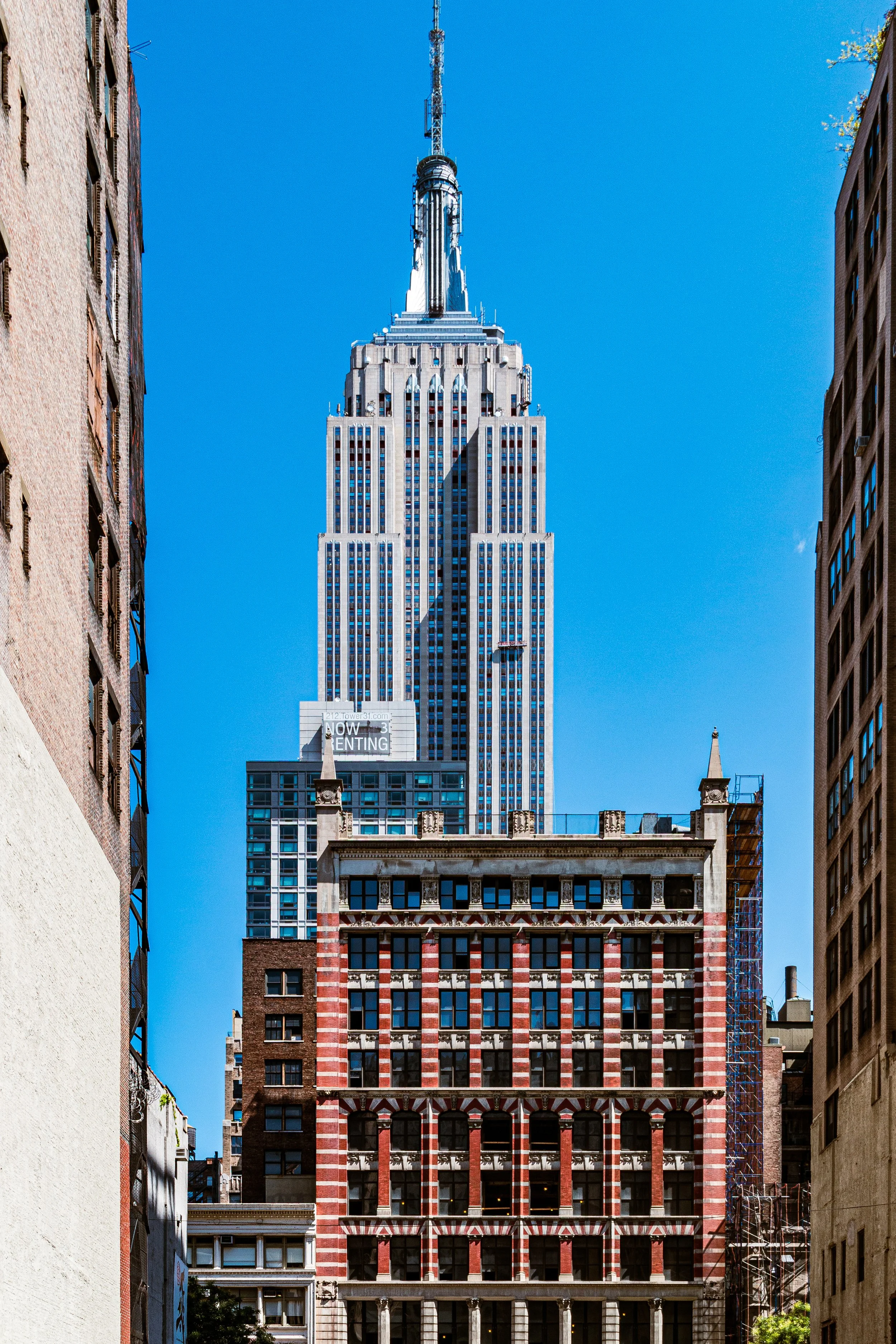 A tall skyscraper, the Empire State Building, in New York City framed by shorter buildings on either side. The blue sky is clear.
