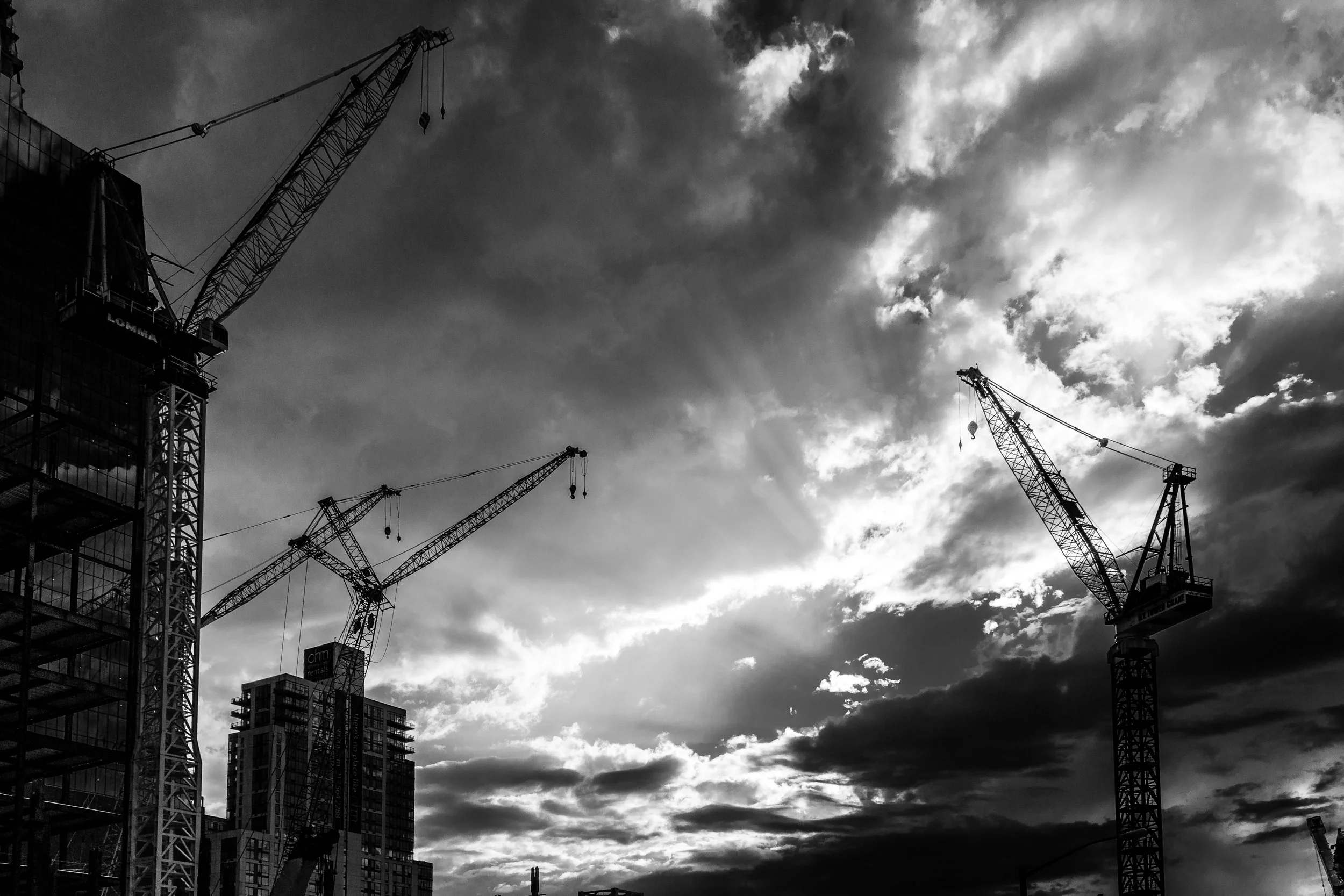 Black and white photo of construction cranes and high-rise buildings against a dramatic cloudy sky.