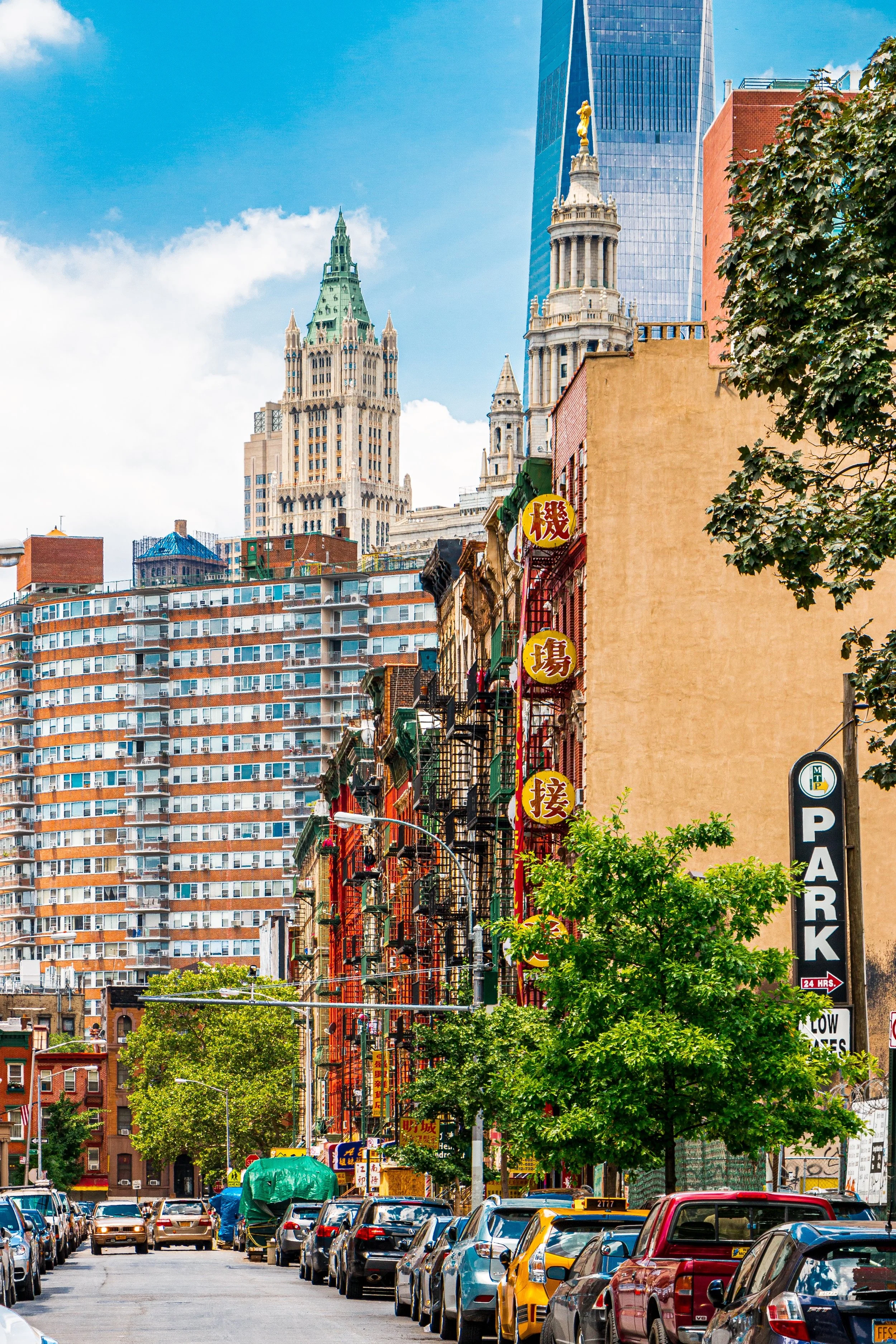 A city street scene with parked cars, trees, and colorful buildings, with tall skyscrapers in the background, including a historic-looking tower and modern glass buildings.