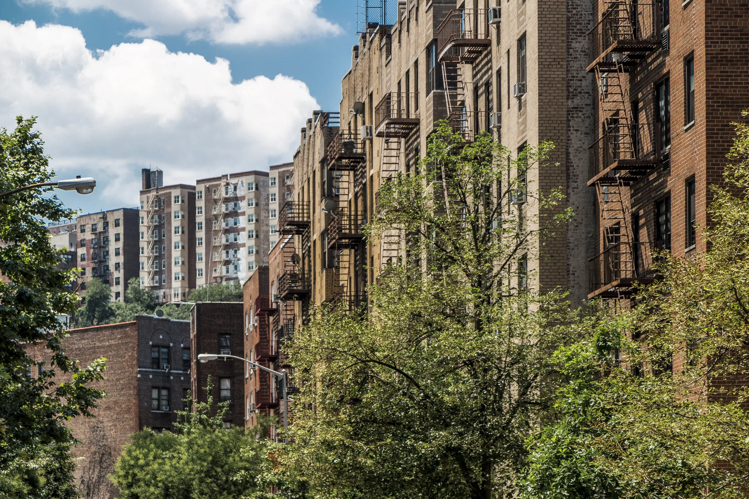 Several multi-story brick apartment buildings with fire escapes on the exterior. Green trees line the street below, and the sky is partly cloudy.