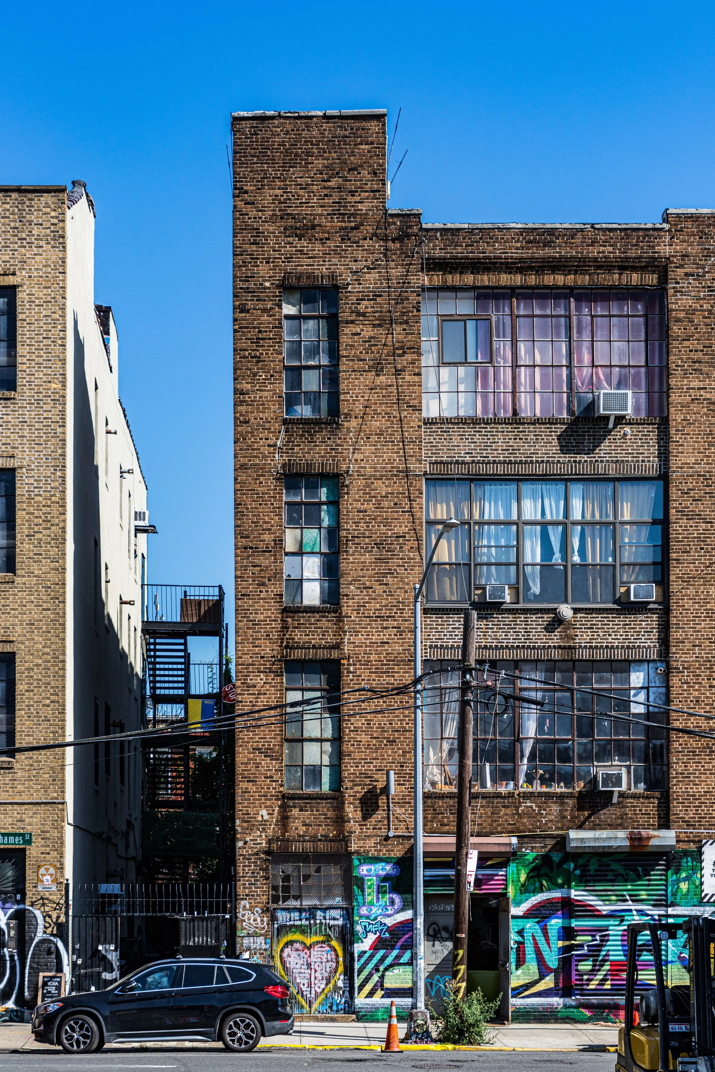 A multi-story brick building with windows, some of which have curtains, air conditioning units, and graffiti on the lower part of the building. A black car is parked on the street in front of it, with street art and graffiti on the wall below. A utility pole with wires is also visible.