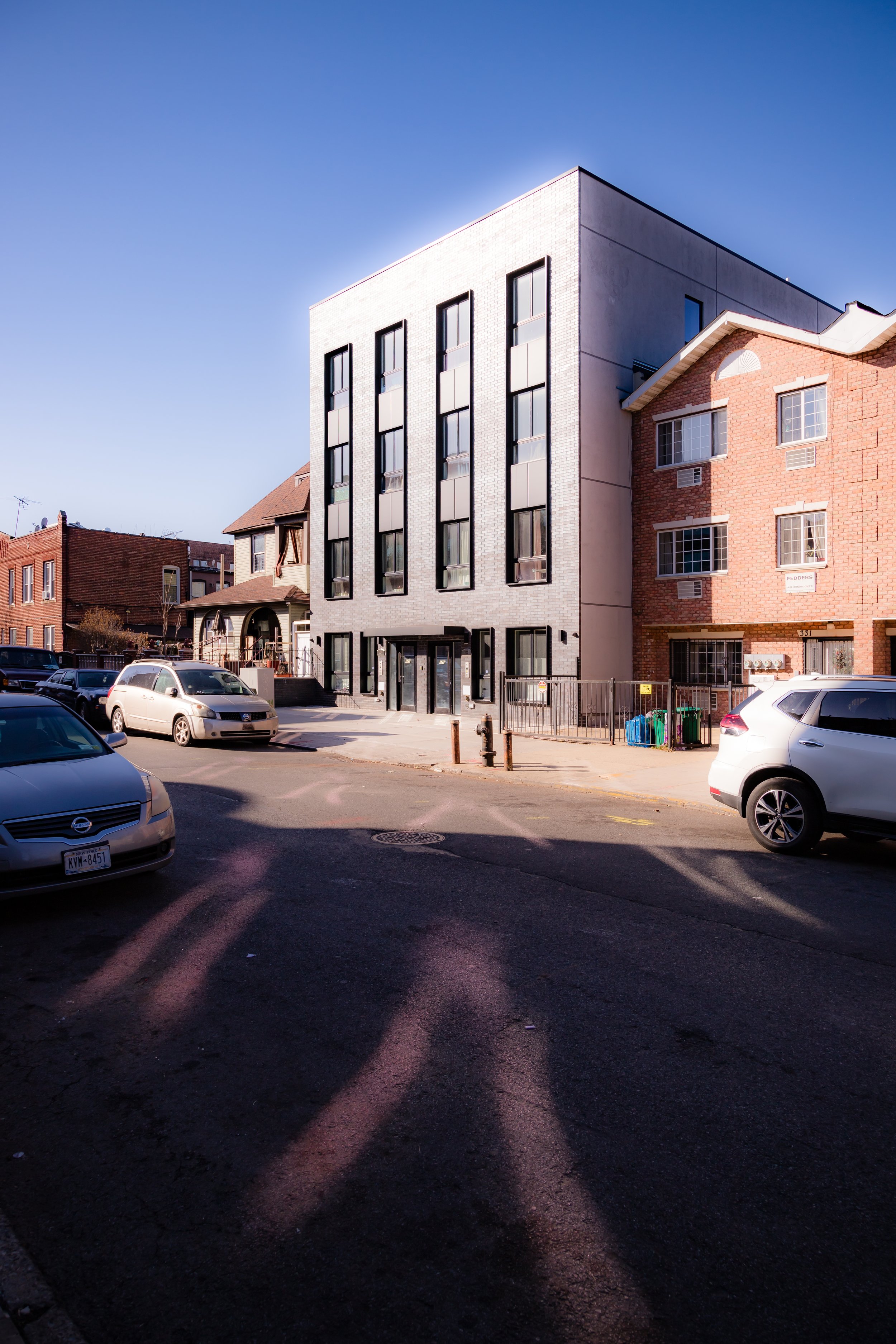 Photo of a city street with parked cars in front of modern and older brick buildings, under a clear blue sky.