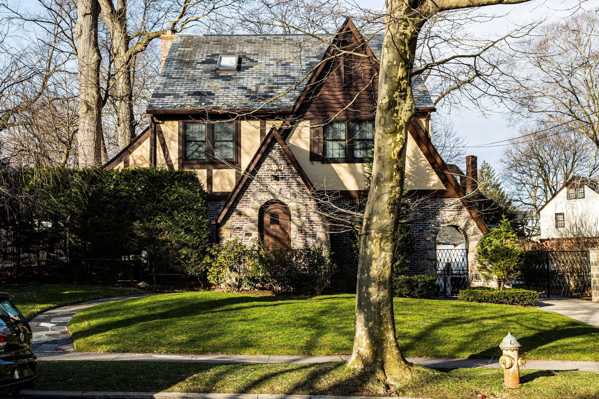 A two-story house with a slate roof, brick and stucco walls, and black window frames, surrounded by trees and a green lawn.