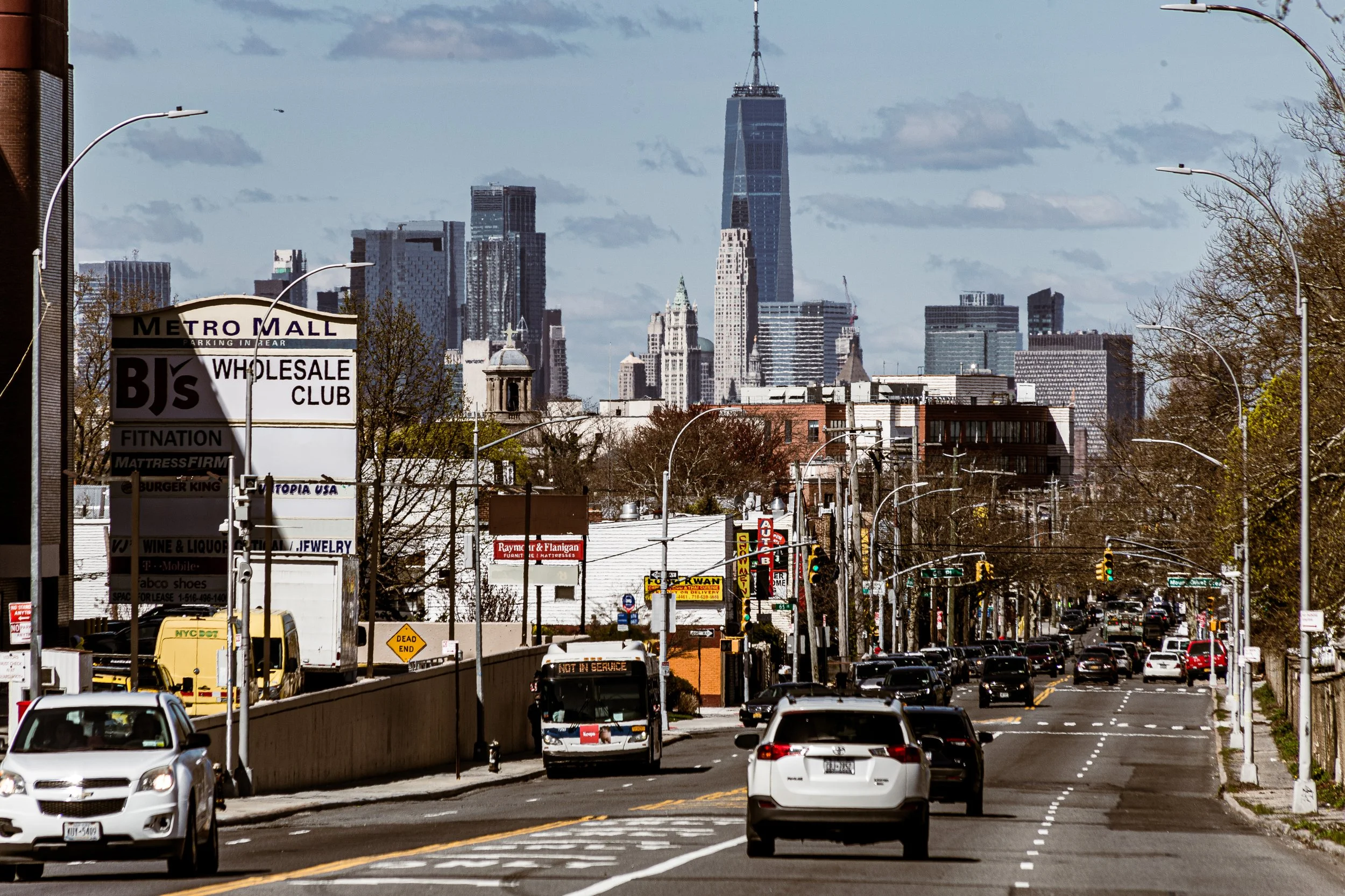 City street with cars driving and parking, advertising signs, and the New York City skyline including the One World Trade Center in the background.