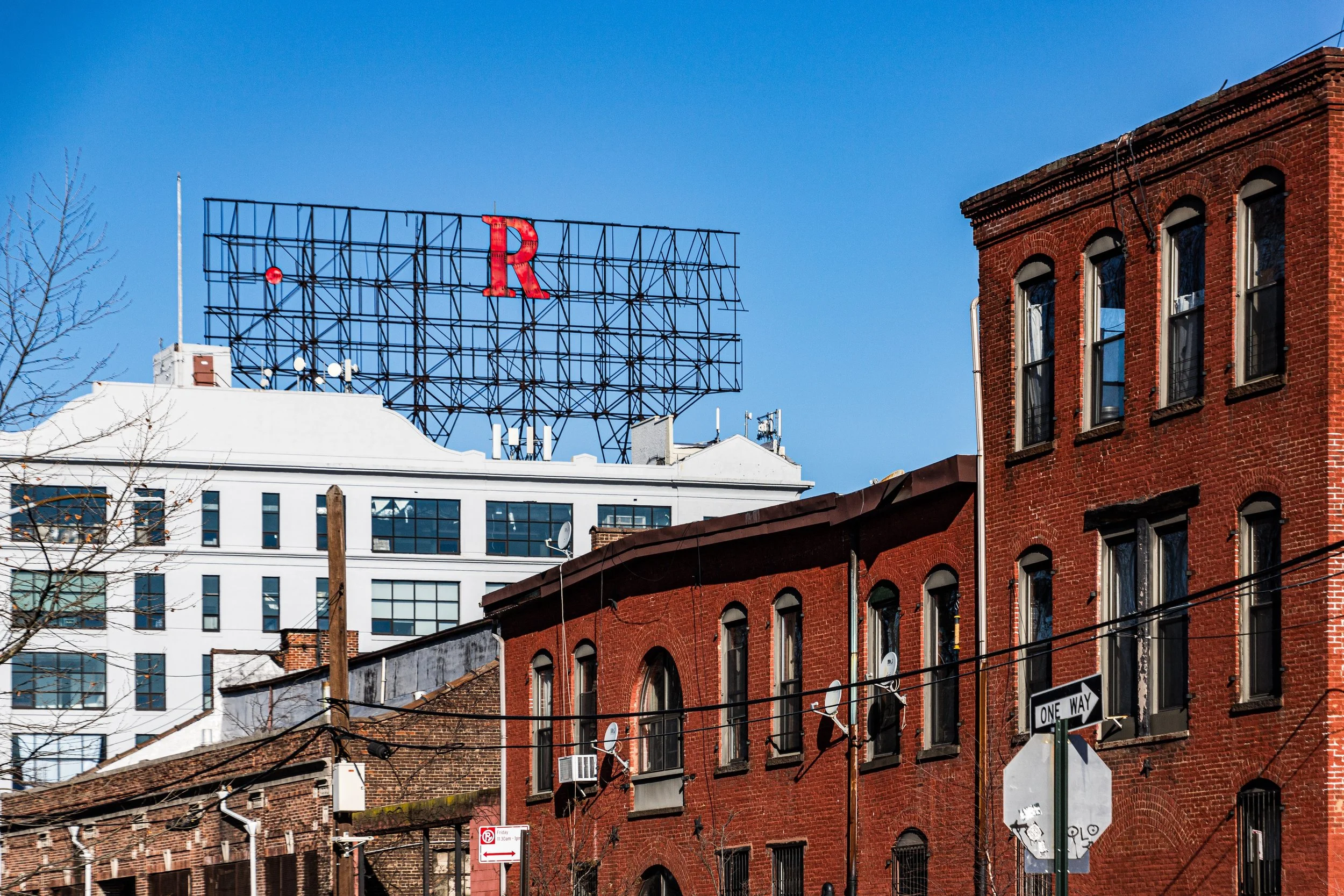 Street view showing red brick buildings, a white building, traffic signs, and a large billboard with a red letter 'R' against a clear blue sky.