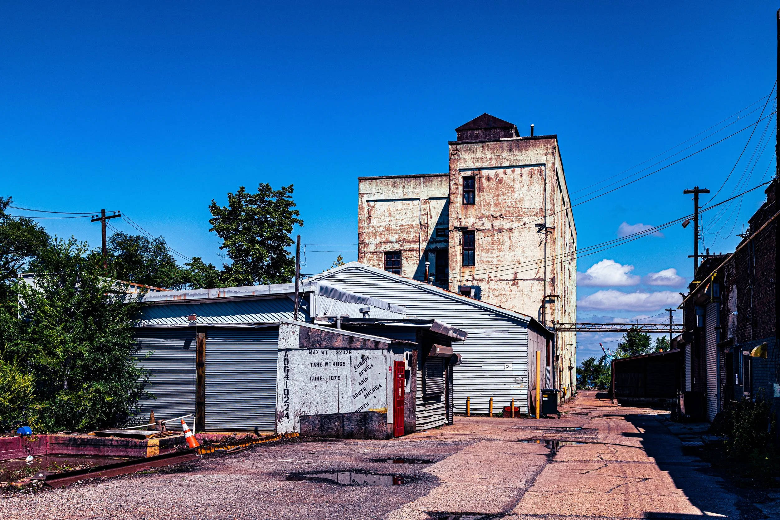 An old industrial building with rusty peeling white paint, surrounded by smaller metal structures and an unpaved street with puddles, under a clear blue sky.