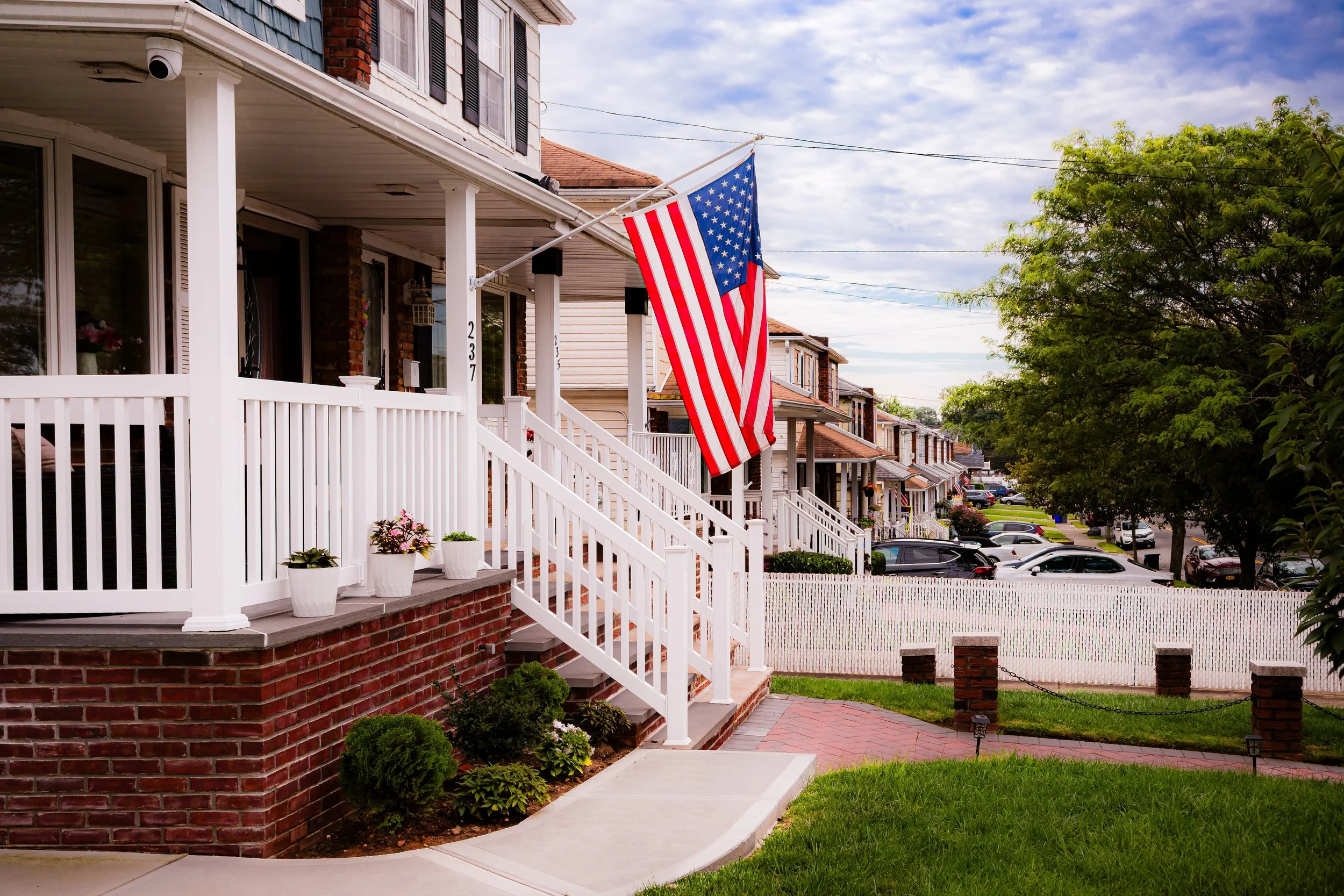 American flag hanging from a porch of a row of brick and white houses, with a white picket fence and a tree-lined street in the background.