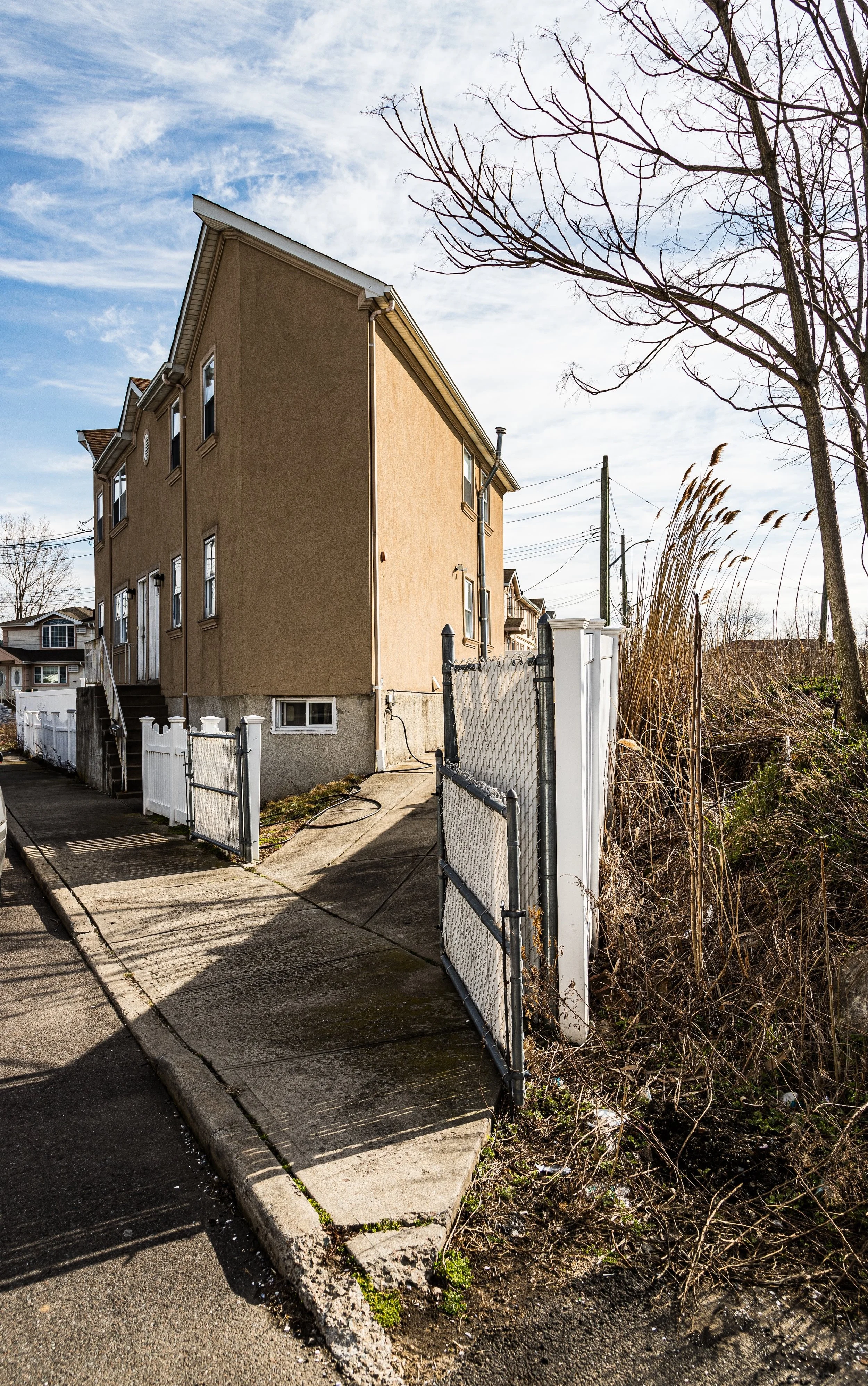 A residential area with a beige multi-story house, a white fence, a sidewalk, and a chain-link gate. Bare trees and tall dry grasses are visible on the right, with a partly cloudy sky overhead.