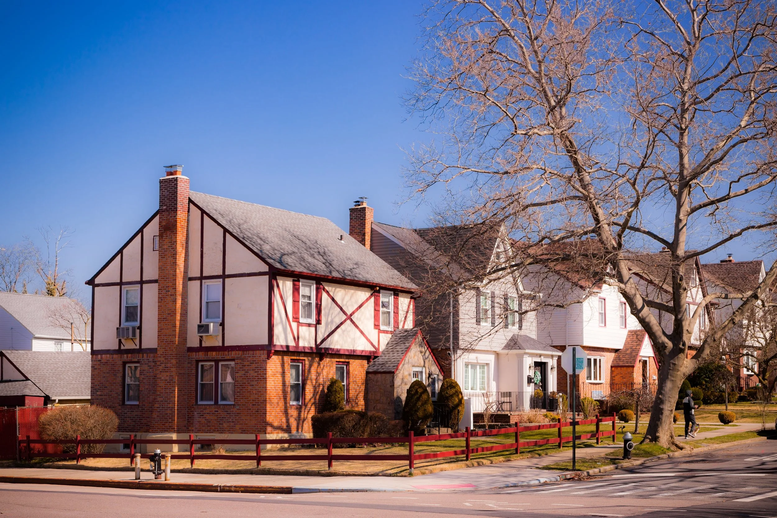 A residential neighborhood with old-style houses, including a two-story house with a brick and stucco exterior, a large leafless tree, sidewalks, and a person walking a dog on a sunny day.