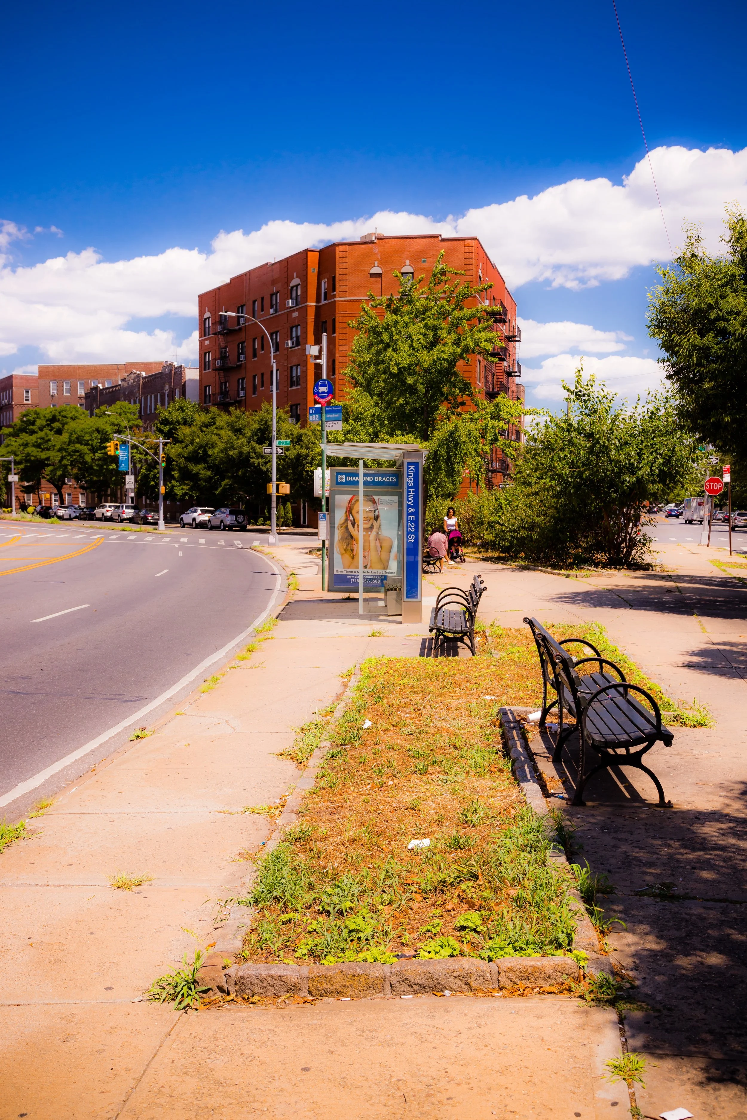 A city street with a bus stop, benches, trees, and a large brick apartment building under a blue sky with white clouds.