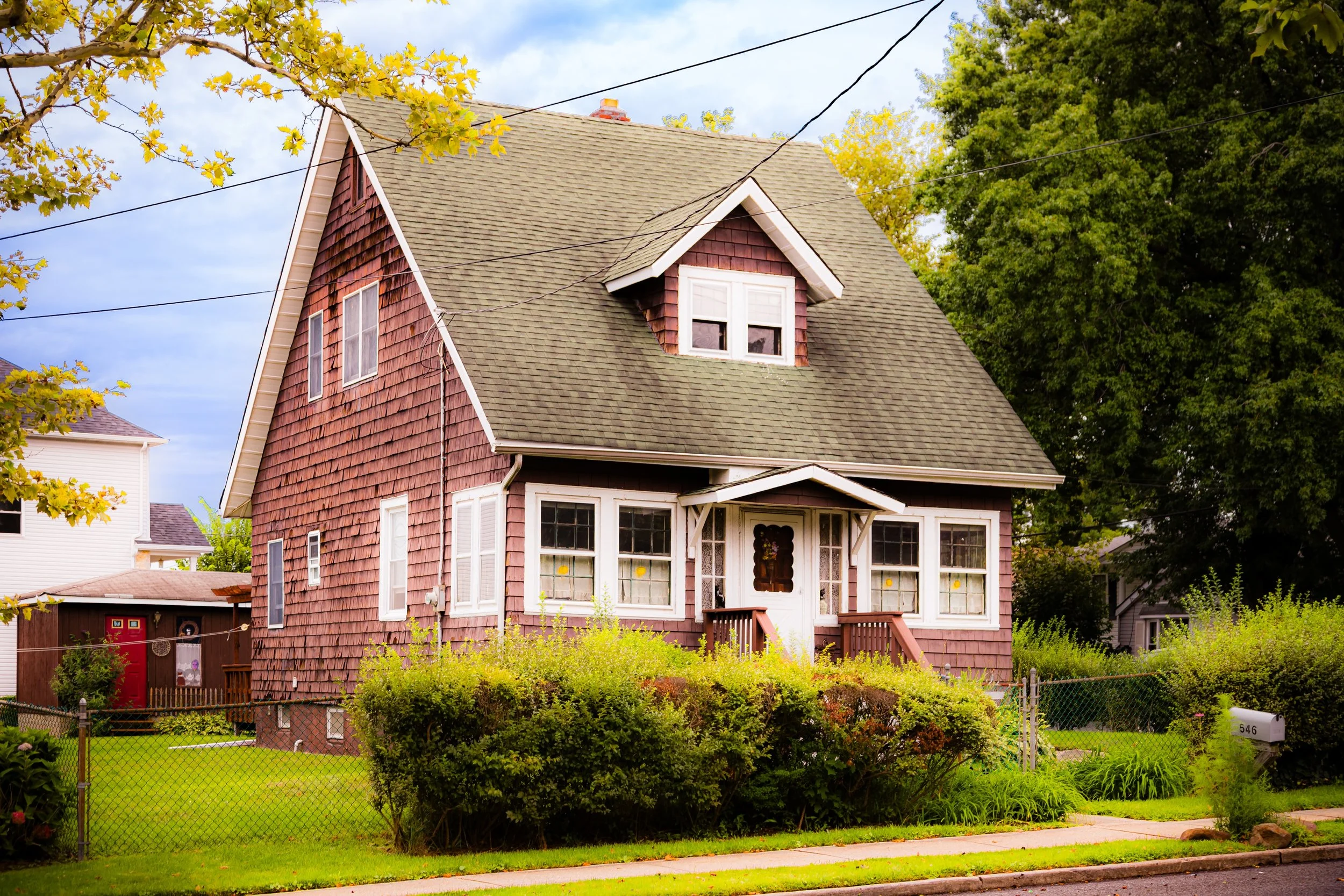 A two-story house with brown shingle siding, a gabled roof, and a small front porch with stairs. The house has multiple white-framed windows and is surrounded by green trees and a well-maintained lawn.