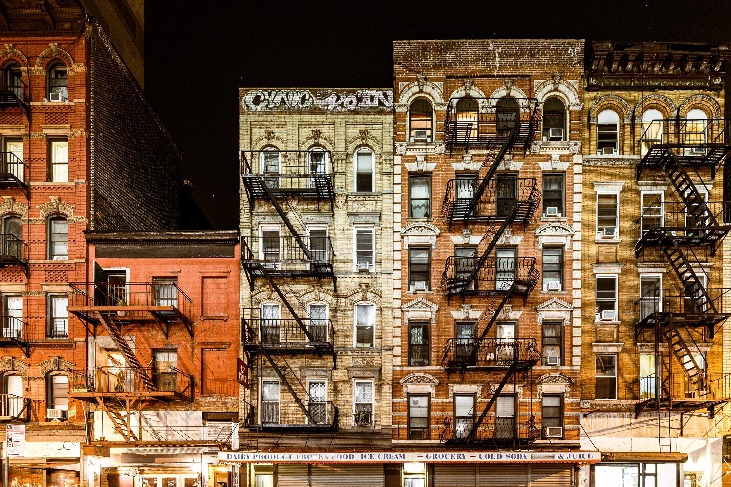 Night view of multi-story brick buildings with fire escape stairs on a city street, storefront with signs for dairy products, frozen foods, ice cream, grocery, cold soda, and juice.