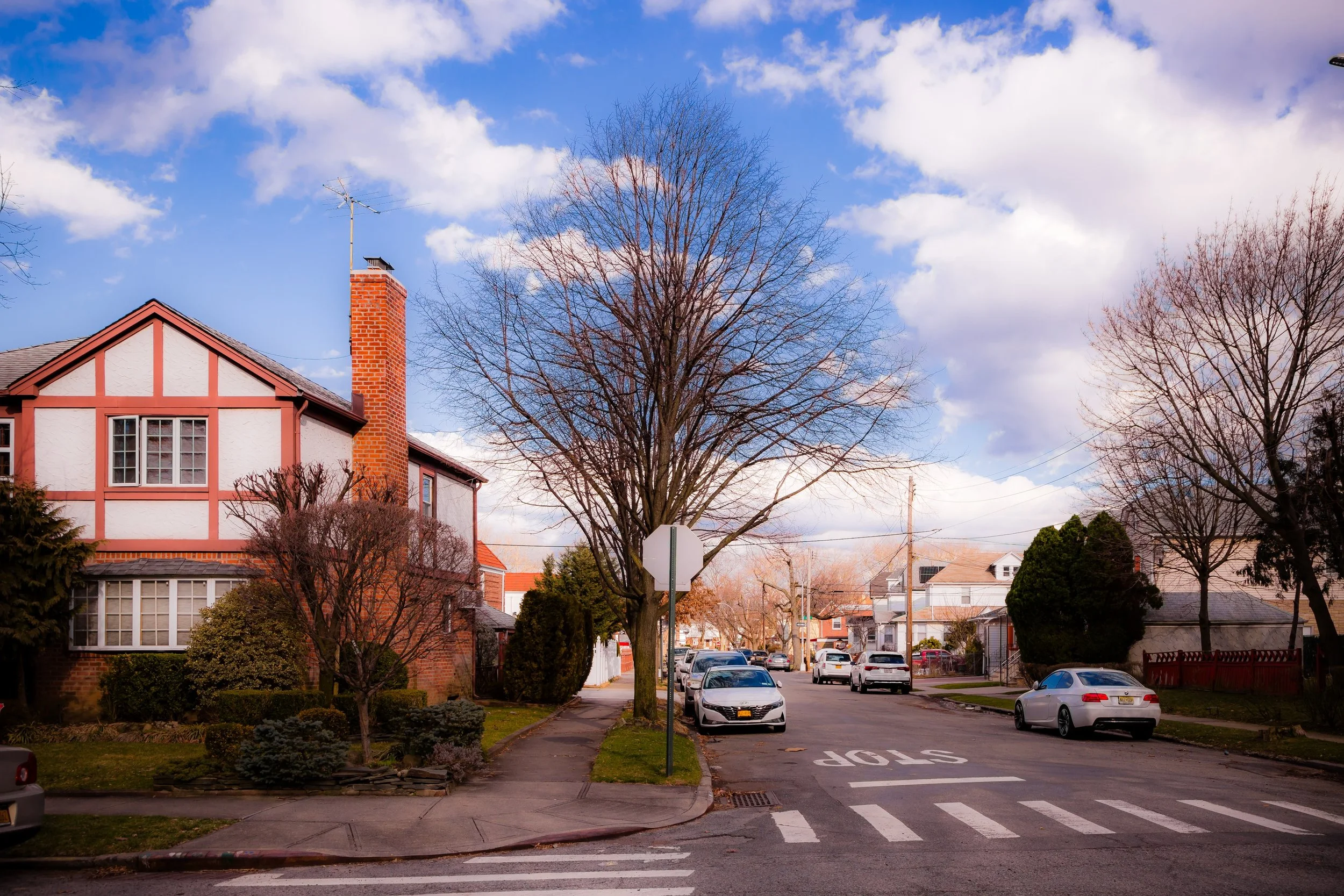 Residential neighborhood street with cars parked along the curb, leafless trees, a stop sign, houses, and a partly cloudy sky.