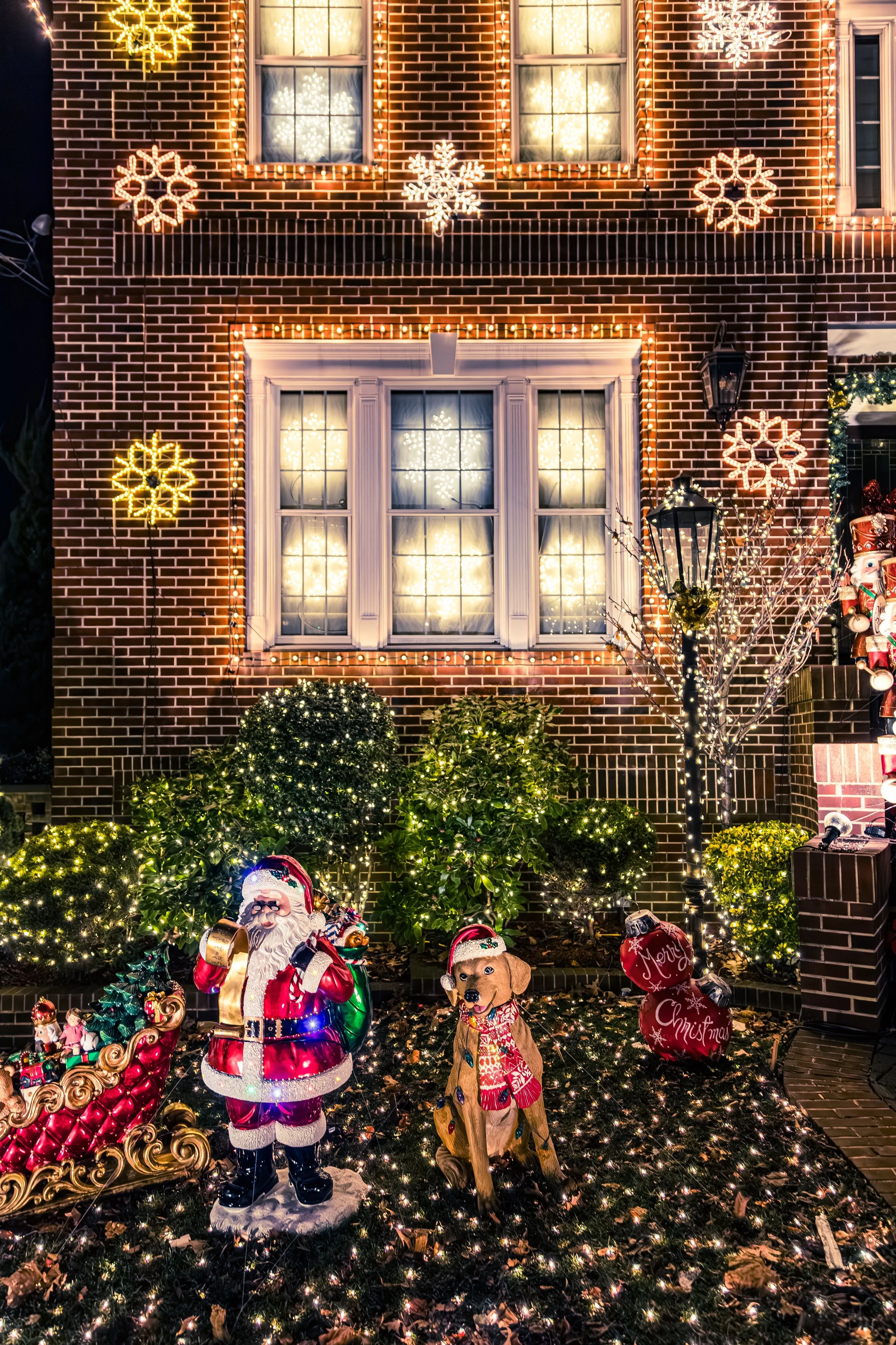 Festively lit front yard with Santa Claus and a dog decorations, illuminated snowflake lights on brick house, and small Christmas trees and bushes decorated with string lights.
