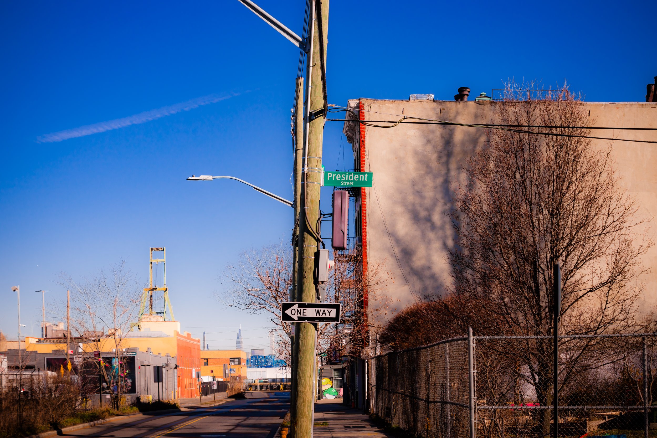 View of a street corner with a green street sign reading 'President Street,' a one-way sign, a utility pole, leafless trees, and a chain-link fence, with colorful buildings and a clear blue sky.