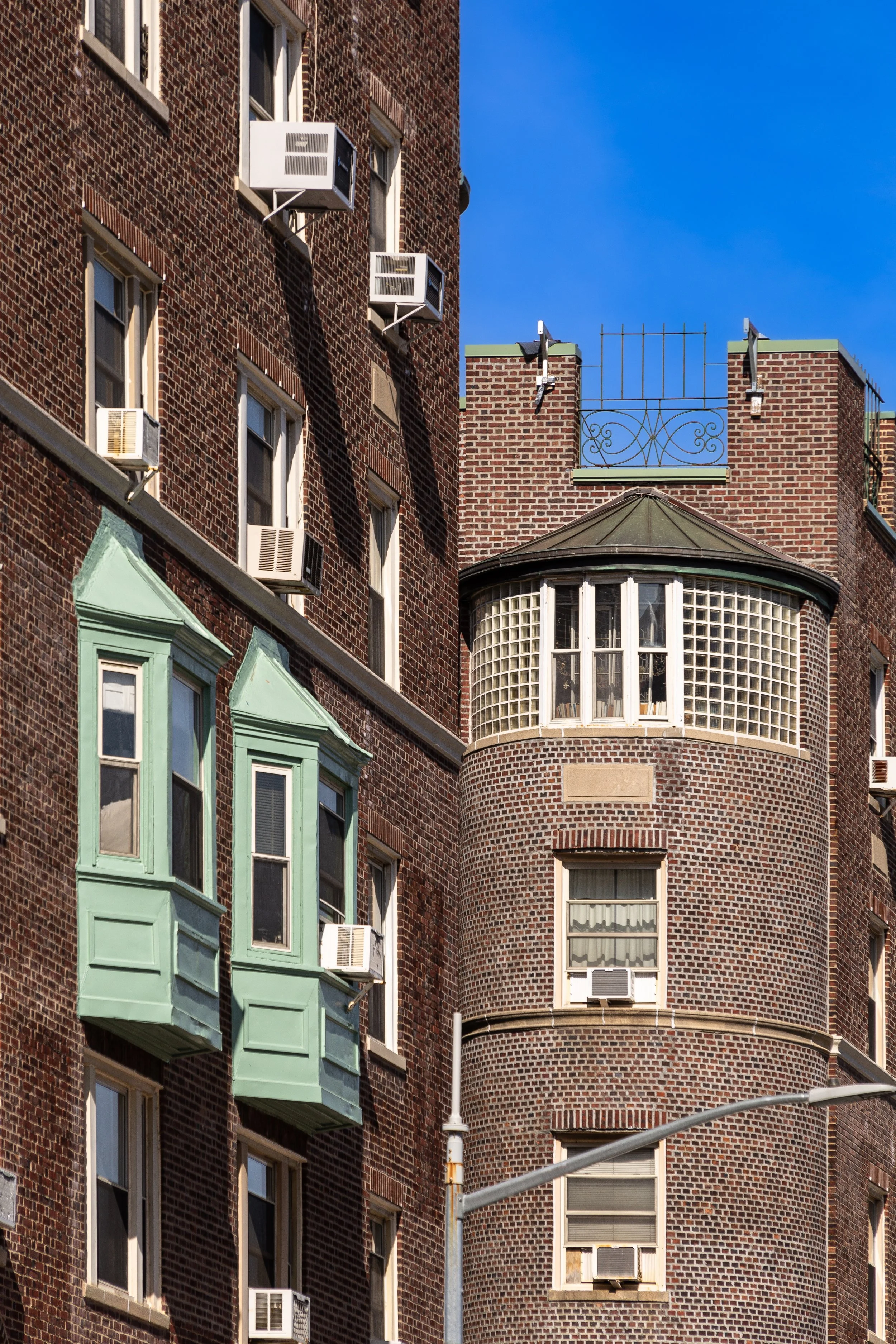 Close-up of a brick apartment building with multiple windows and air conditioning units. Some windows have green bay windows. The building features a rounded corner with glass block windows and a decorative wrought iron structure on the roof. Bright blue sky in the background.