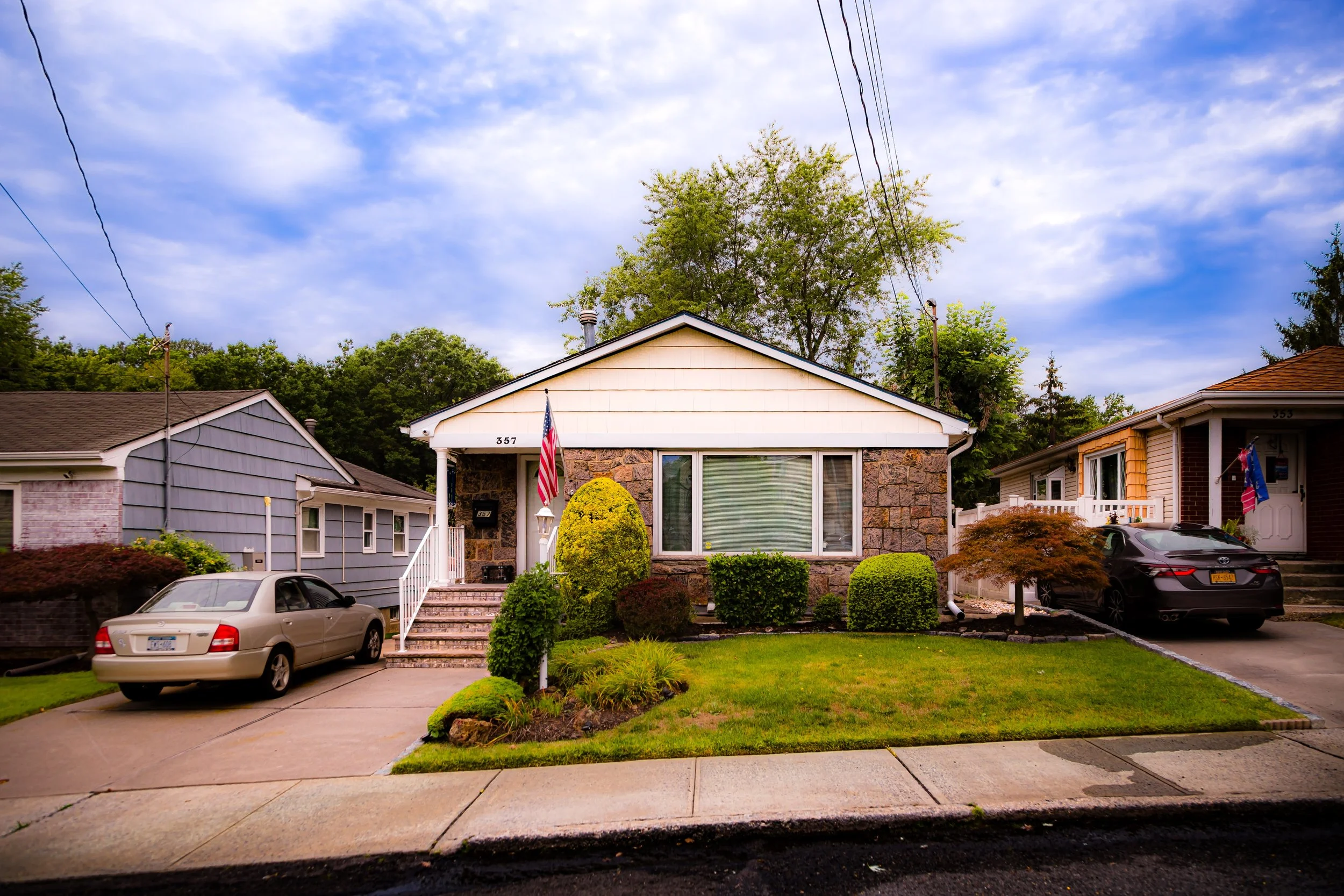 A single-story house with a stone exterior and white siding, front yard with green grass and shrubs, American flags on the porch, cars parked in driveway and on the street, neighboring houses on either side, under a partly cloudy sky.