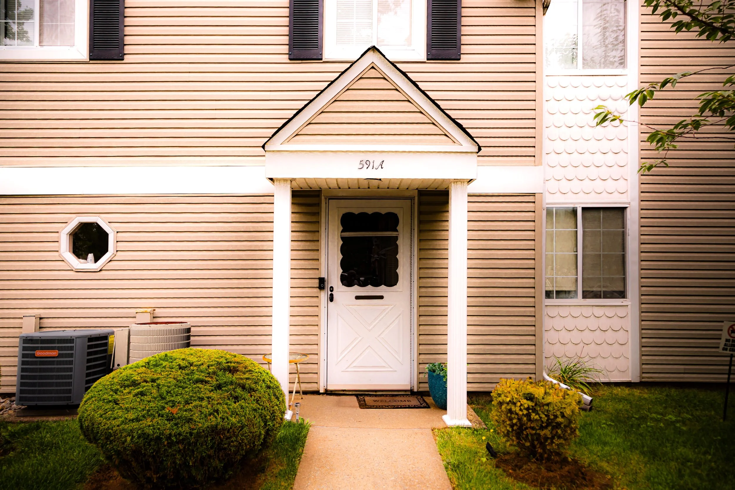 Front entrance of a beige house with a white door, small porch, and a triangular roof. A bush and potted plant are in the yard, with an air conditioning unit visible on the left.