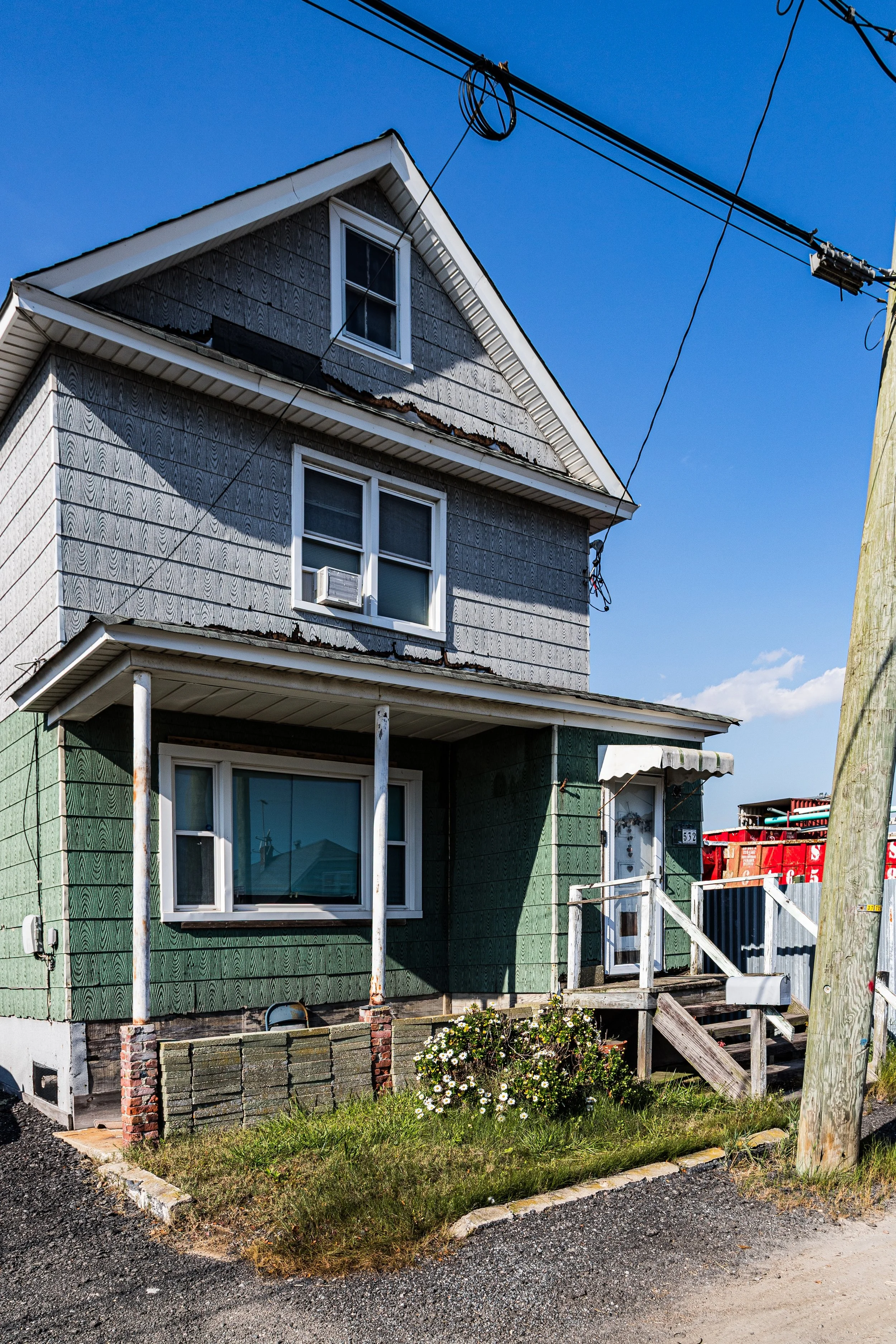 A two-story house with weathered green and gray wooden siding, a small front porch with stairs, and a flowering bush in front. The house has a pitched roof, multiple windows, and power lines overhead against a clear blue sky.
