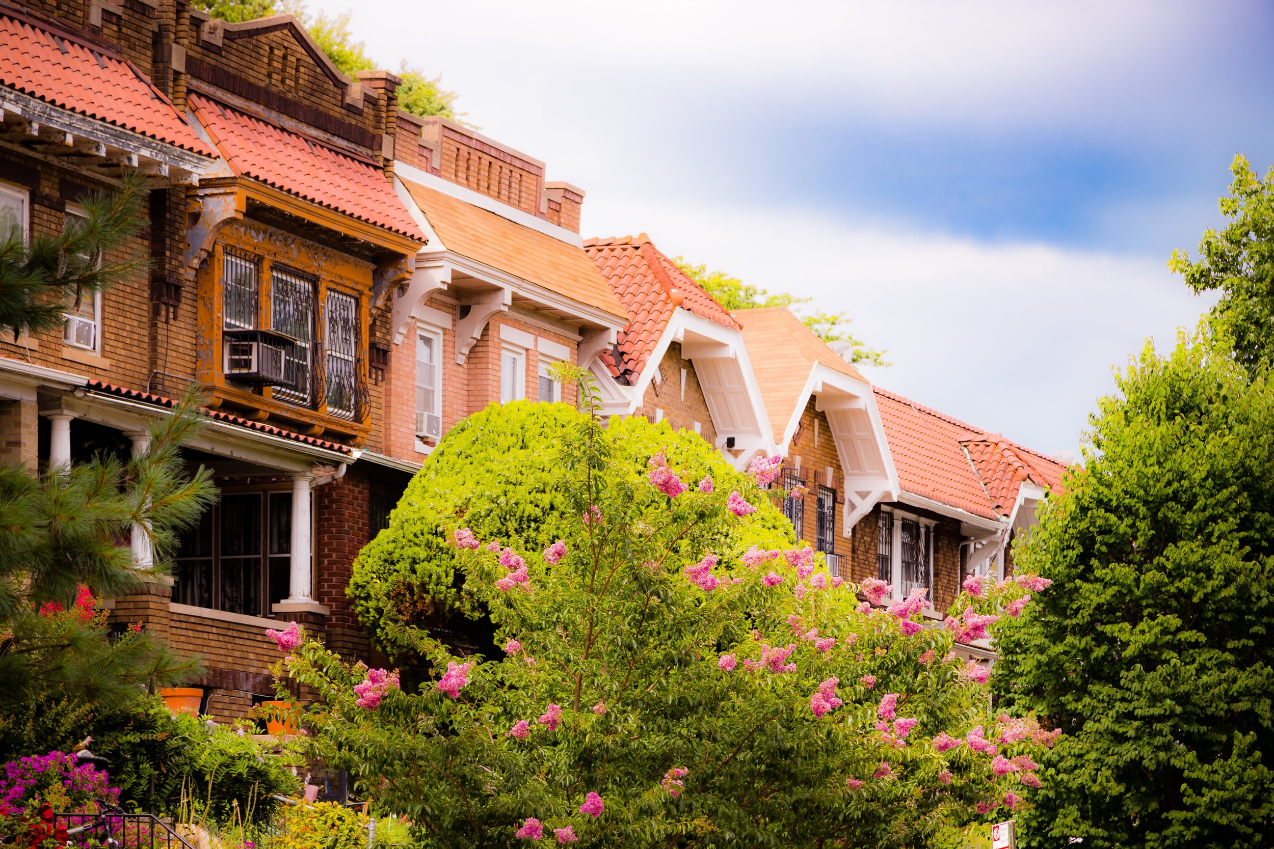 A row of brick houses with red-tiled roofs and white accents, surrounded by green trees and pink flowering bushes.