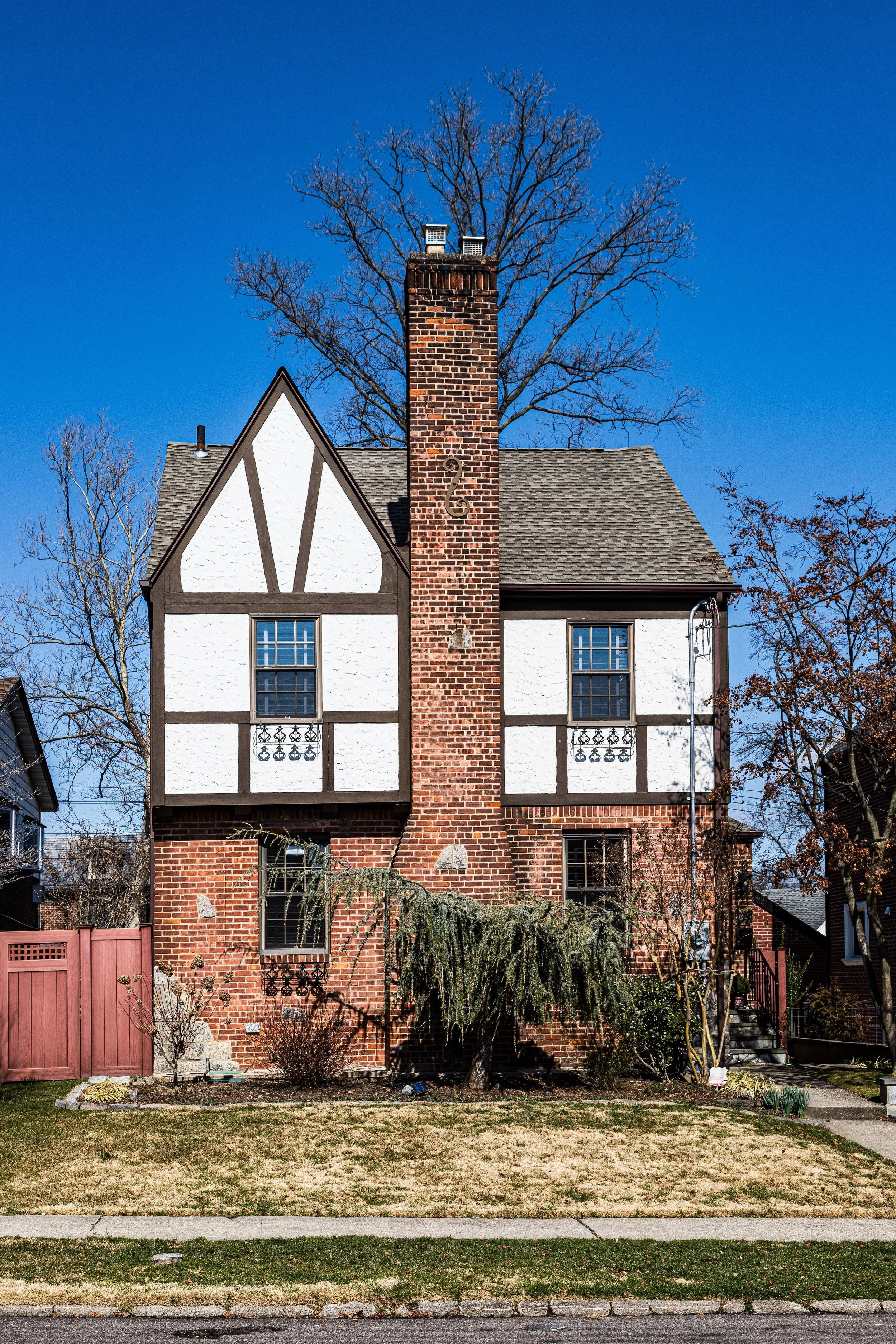 A two-story house with a brick base, white and brown Tudor-style upper floor, and a tall brick chimney, set against a blue sky with leafless trees in the background.