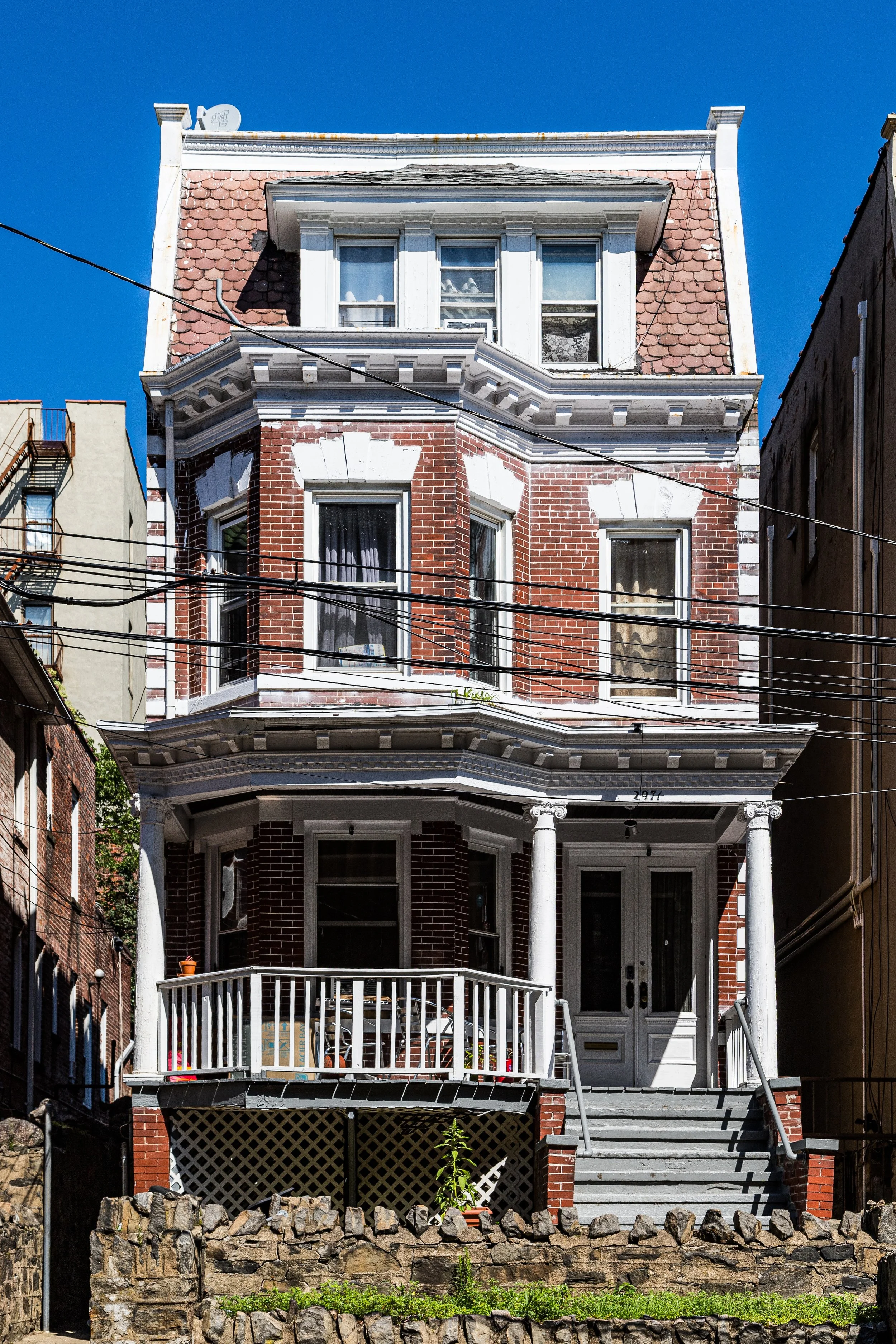 A three-story Victorian-style brick house with white trim, decorative columns at the porch, and multiple bay windows. The house has a small front porch with a white railing and steps leading up to the front door. The sky is clear and blue.