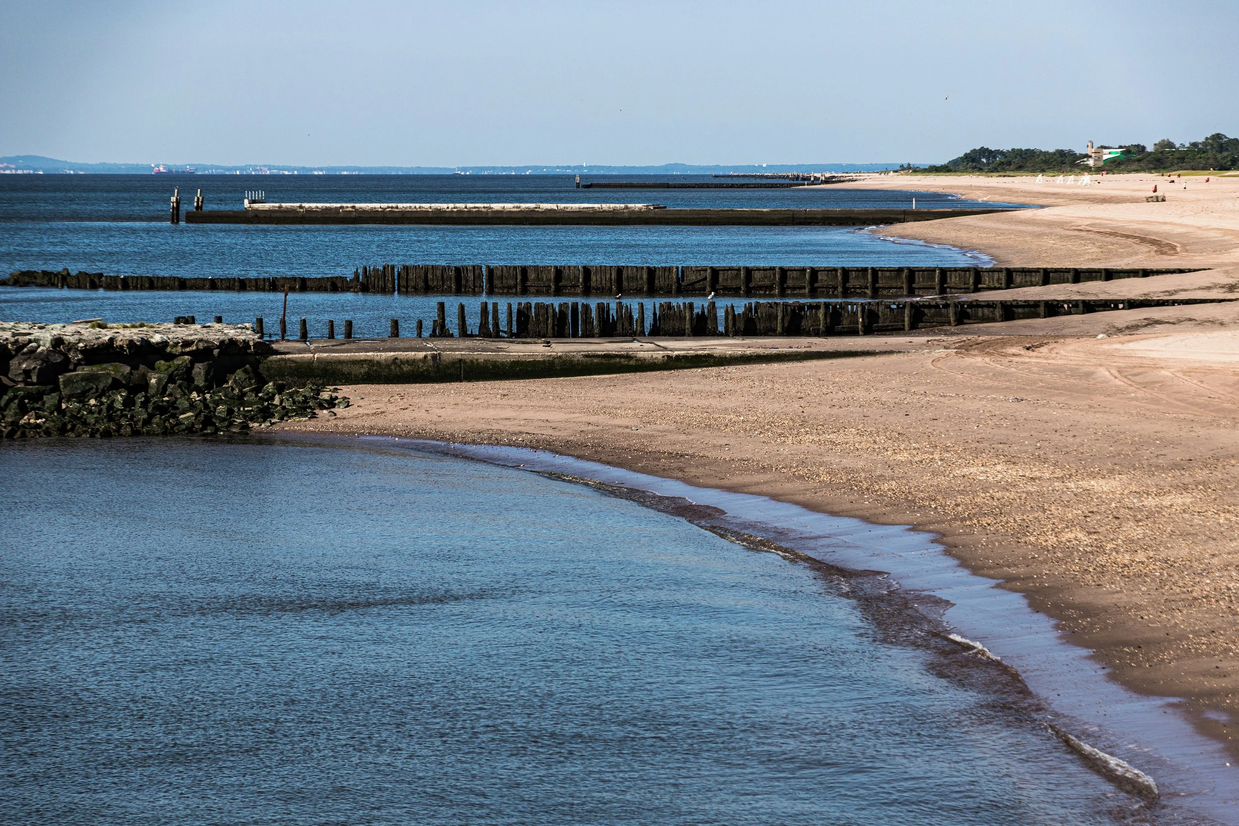 A sandy beach with a calm waterway in the foreground and a larger body of water in the background, separated by wooden breakwaters and a stone wall, under a clear blue sky.