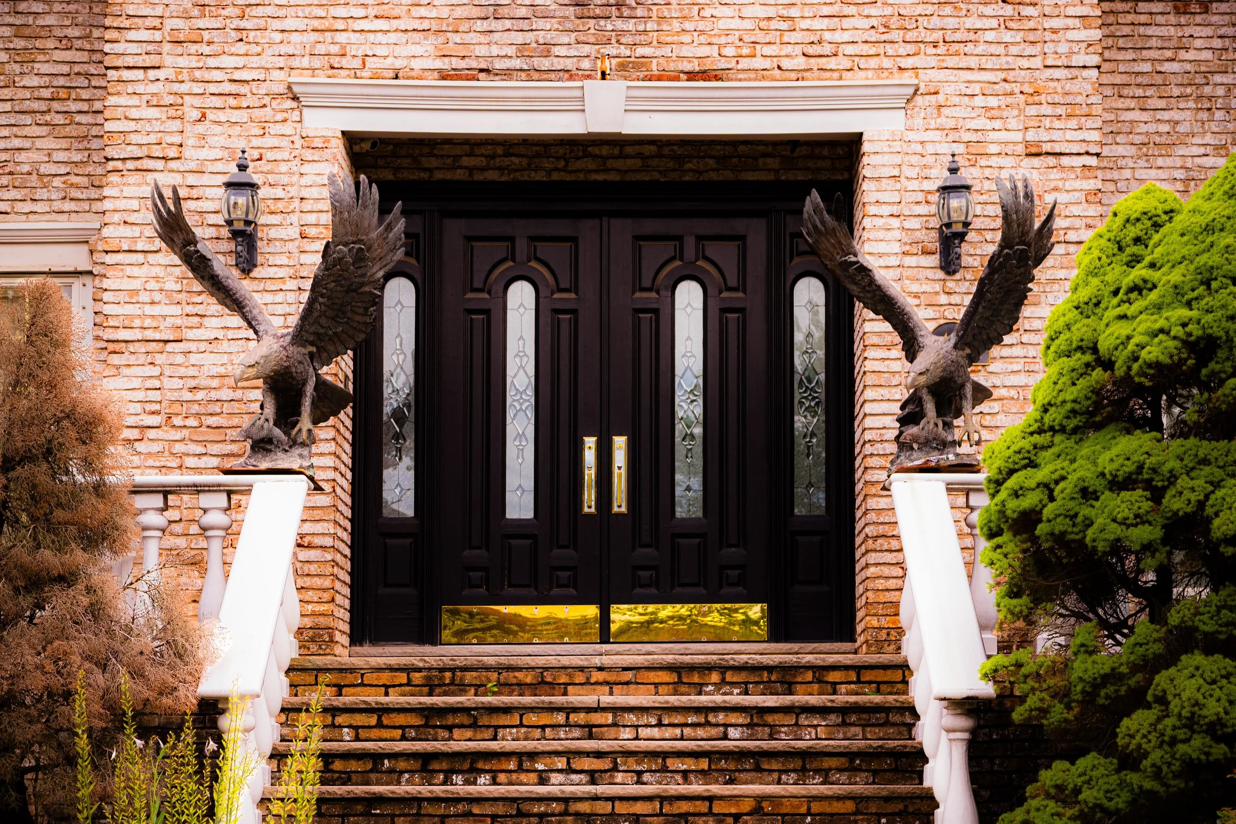Decorative front door with black double doors, glass panels, and brass handle, flanked by two eagle statues on white pedestals, with brick steps, brick wall, and lush green bushes.