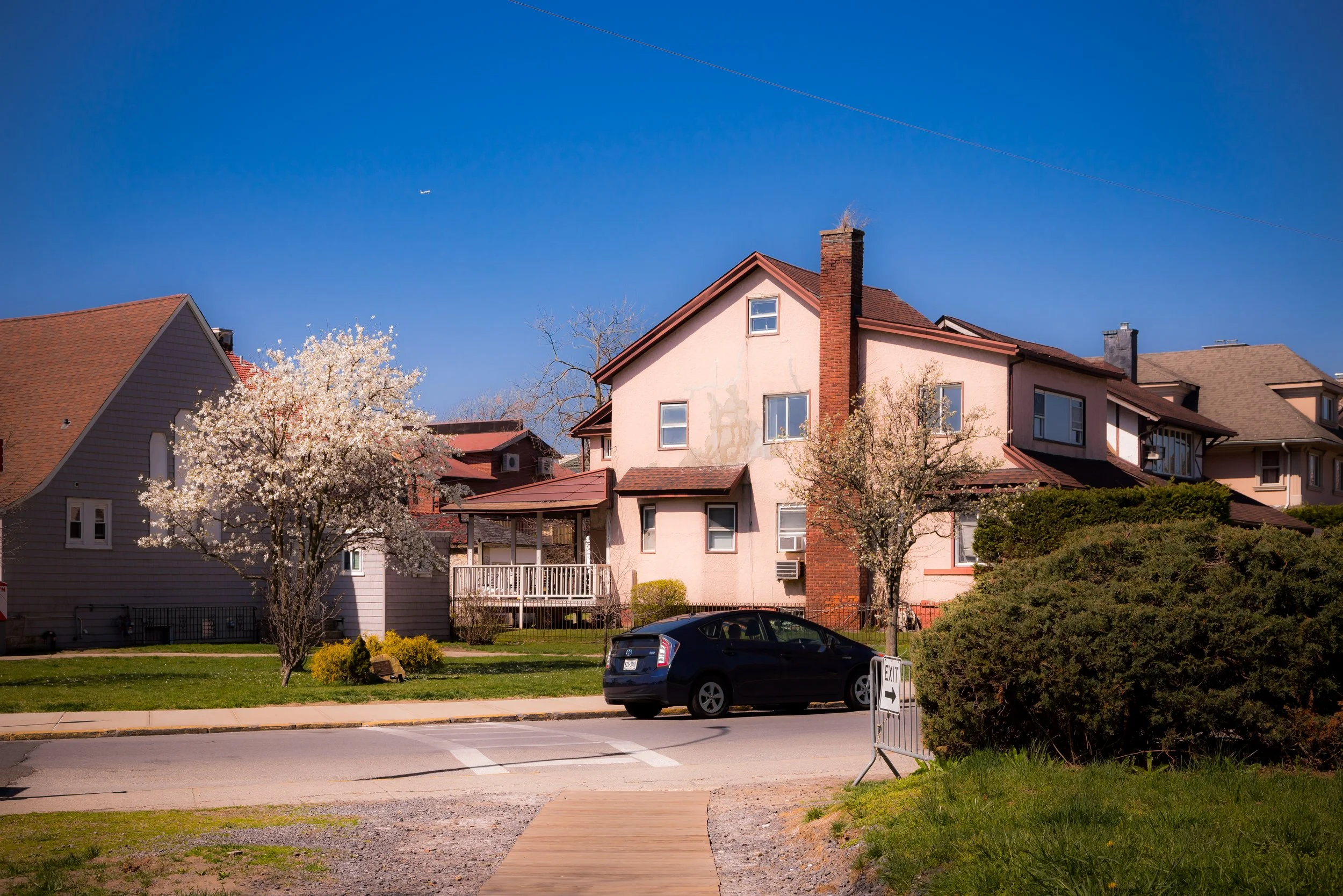 A residential neighborhood with a pink multi-story house, a blooming tree, a parked black car, and a blue sky with a plane flying overhead.