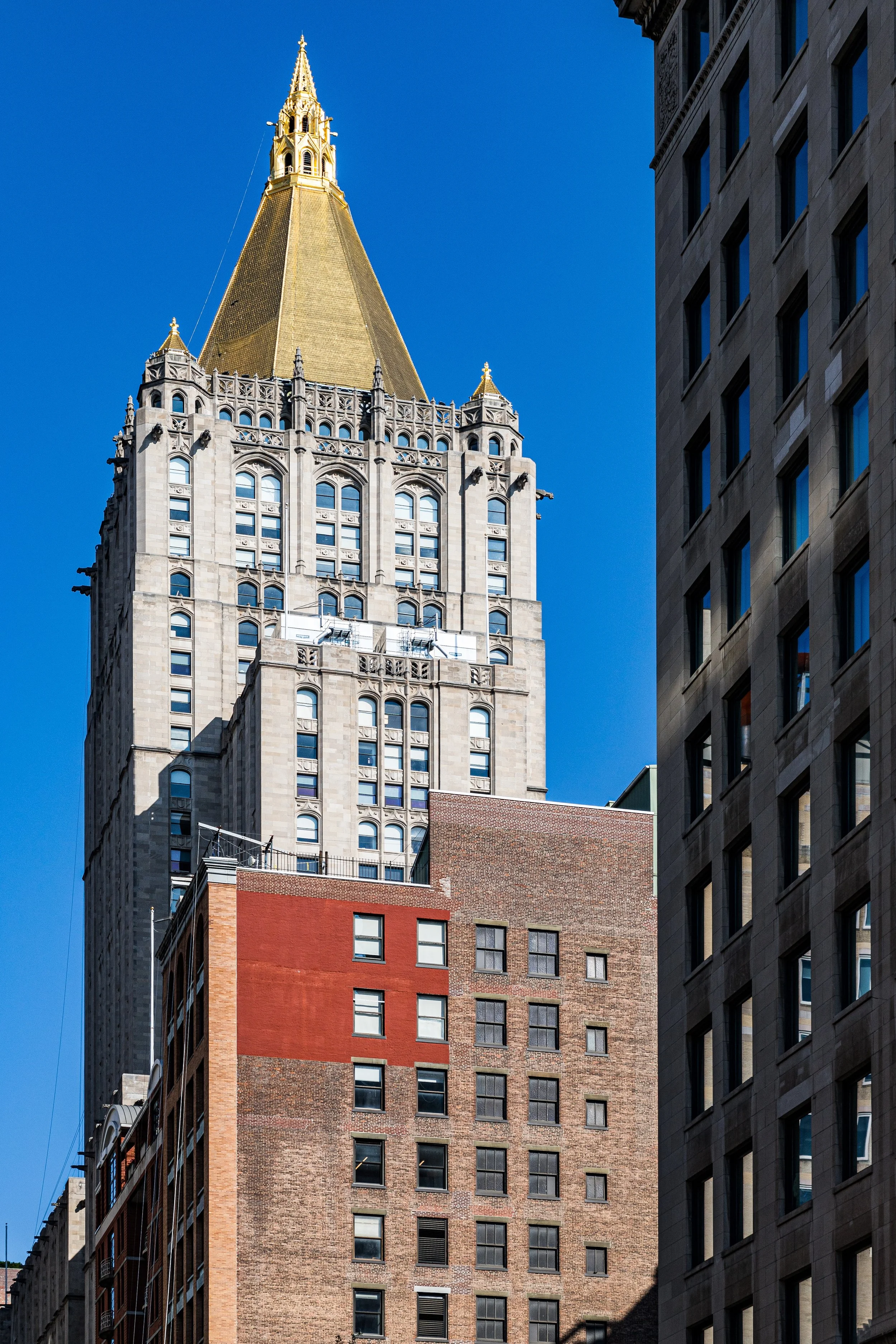 View of a tall building with a golden spire against a clear blue sky, surrounded by shorter buildings in an urban cityscape.