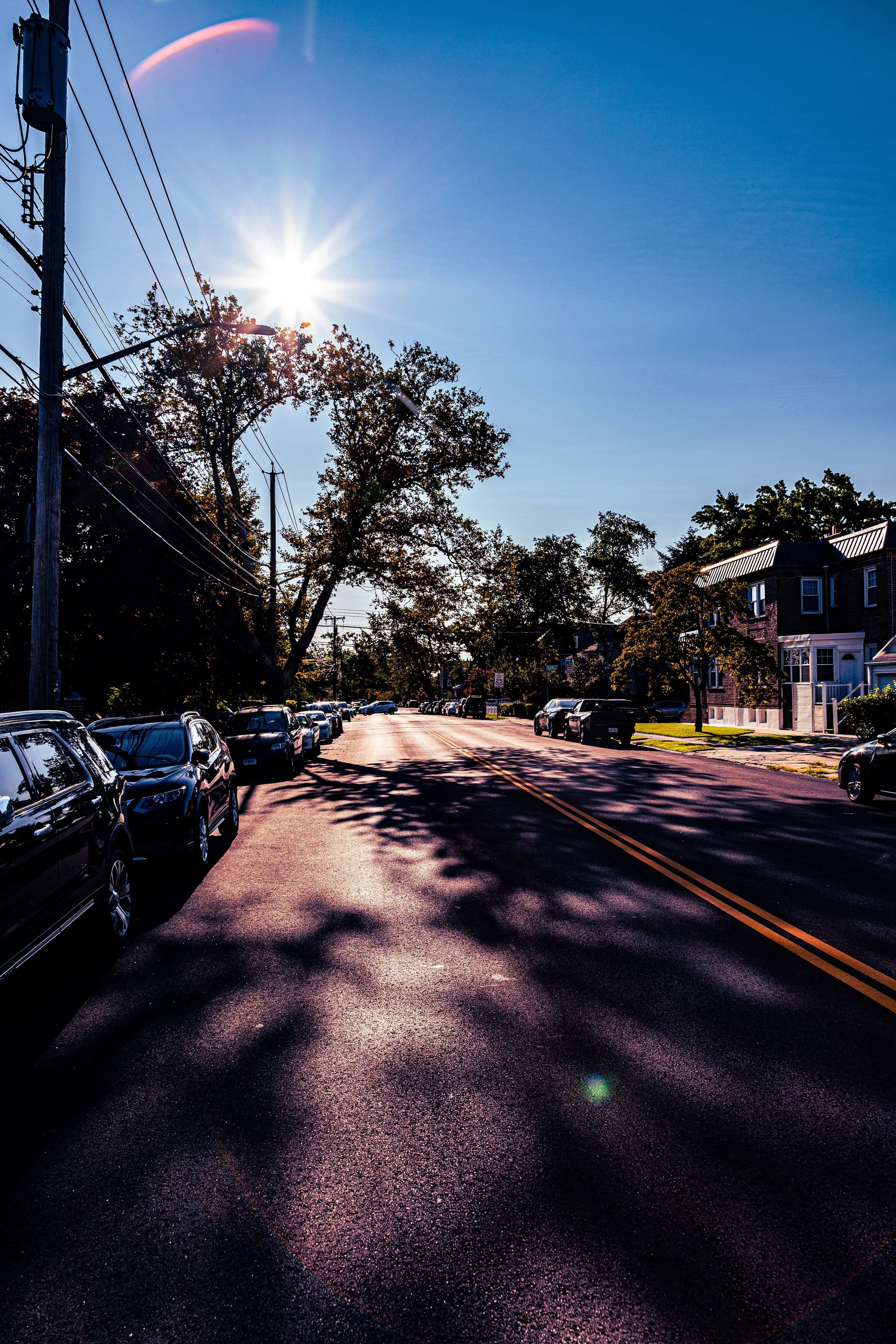 Sun shining brightly over a residential street with parked cars and trees casting shadows during daytime.