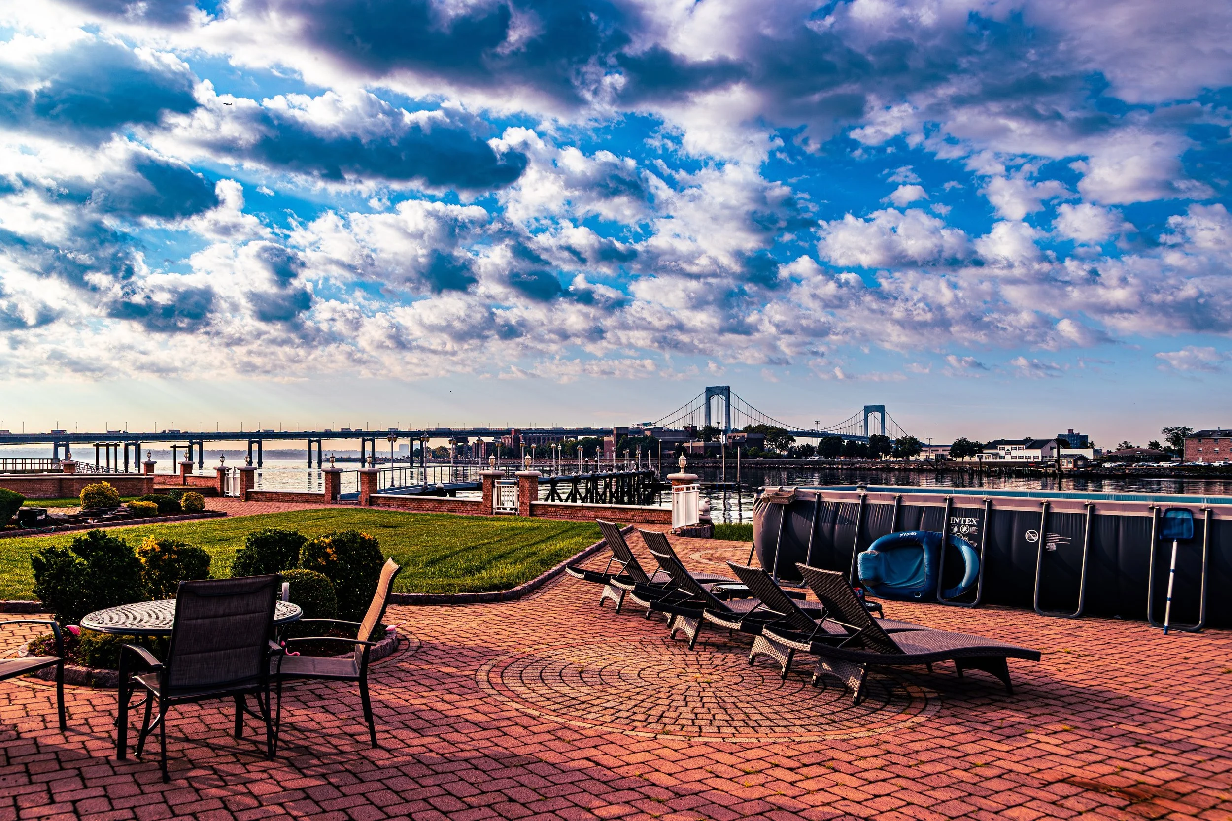 View of a waterfront patio with lounge chairs, a small patio table with chairs, a green lawn, a bridge in the background, and a cloudy sky.