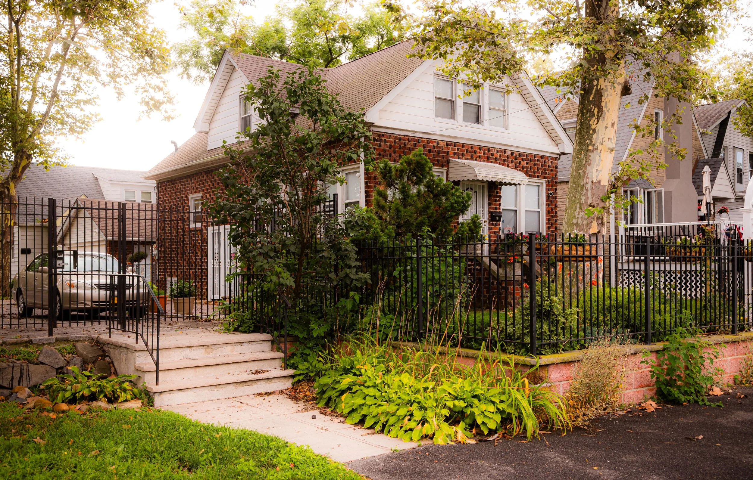 A two-story house with brick on the front and white siding on the upper part, surrounded by a black metal fence and green plants, with a driveway and a car parked on the left side.