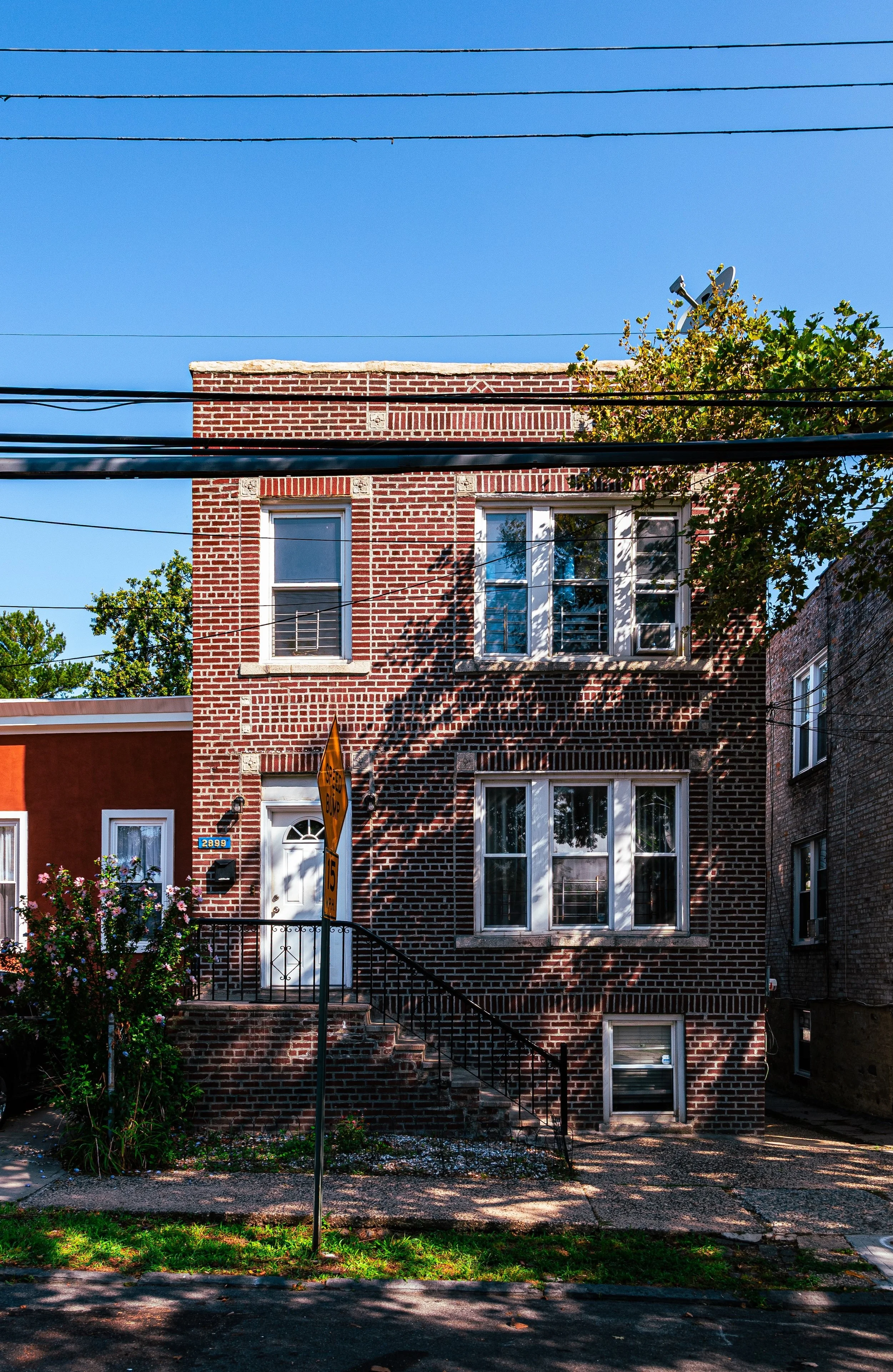 A three-story brick house with white-framed windows and a white front door, located on a residential street with trees and overhead power lines, under a clear blue sky.
