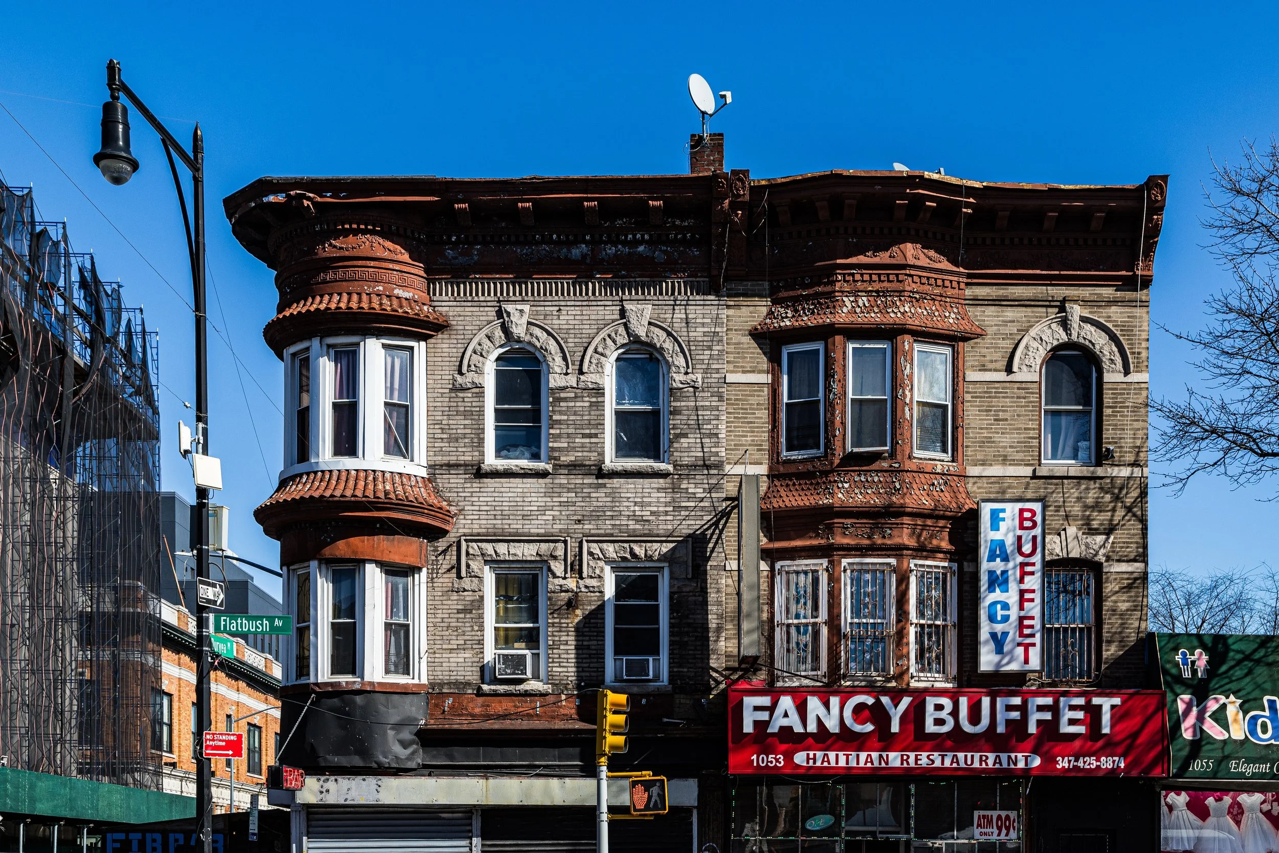 A three-story brick building with bay windows on the top floors, featuring an orange terracotta roof and decorative architecture. The ground floor has signage for 'Fancy Buffet,' a Haitian restaurant, with a vertical signboard and a red awning. A street sign reading 'Flatbush Ave' is visible, along with a traffic light in front.