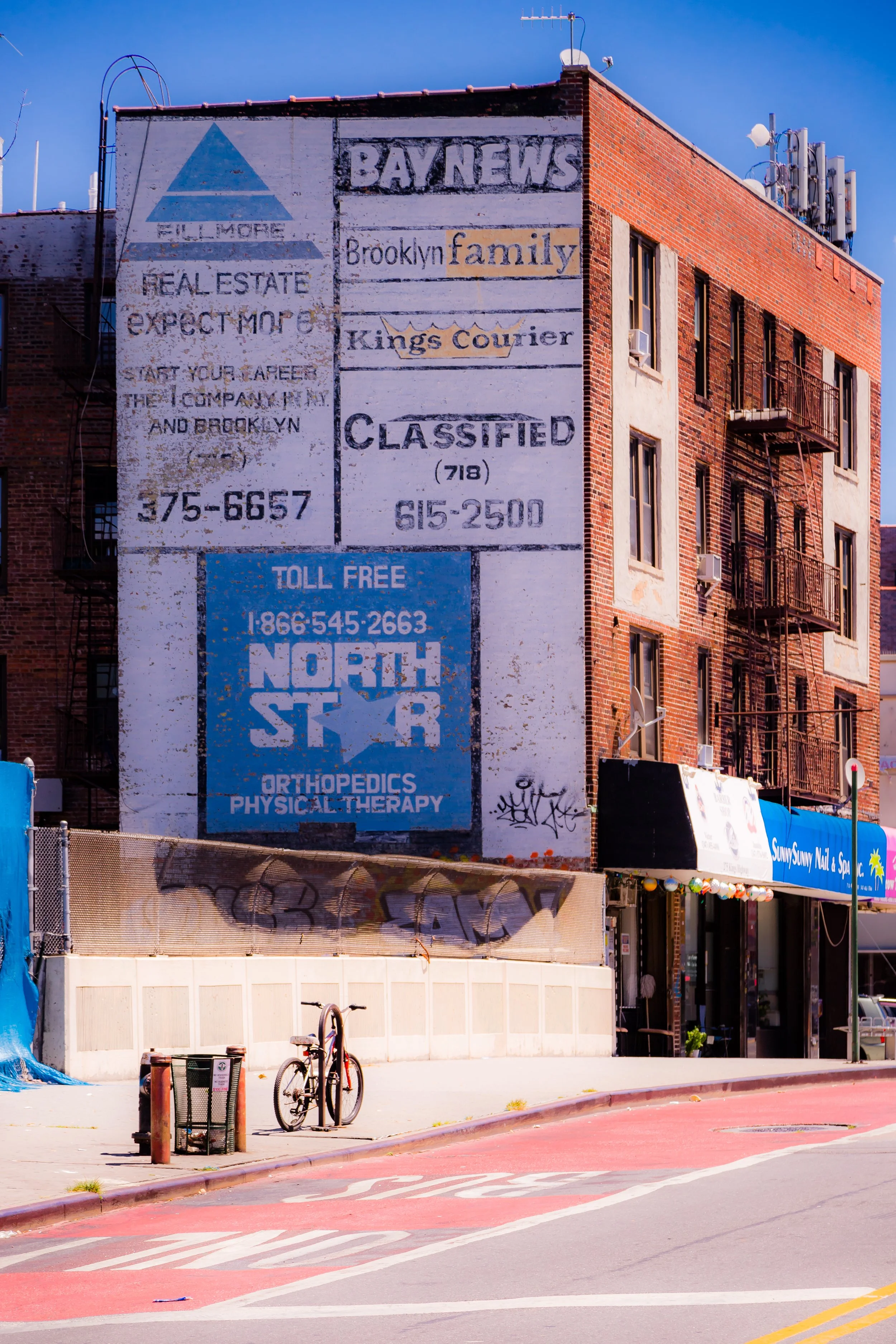 A multi-story brick building with various signs painted on the side, including a large blue and white sign for North Star Orthopedics and Physical Therapy, and signs for Brooklyn Family, Kings Courier, Bay News, and a classified ad. The building has fire escapes and air conditioning units on the windows. There is a bicycle locked to a pole and graffiti on a lower wall. The street has a red bike lane and a store with a blue awning.