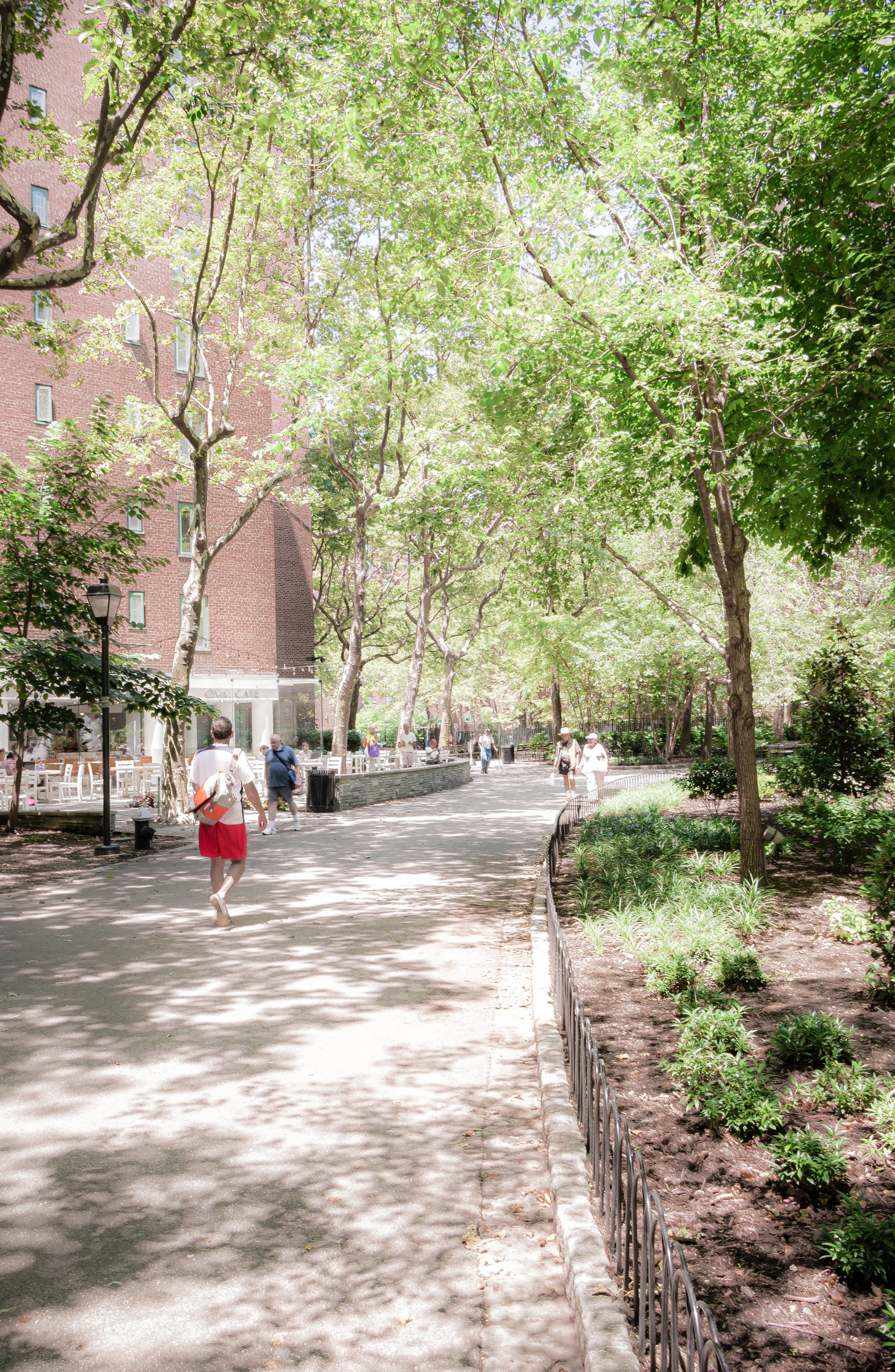 People walking along a shaded park pathway with trees and greenery on a sunny day.