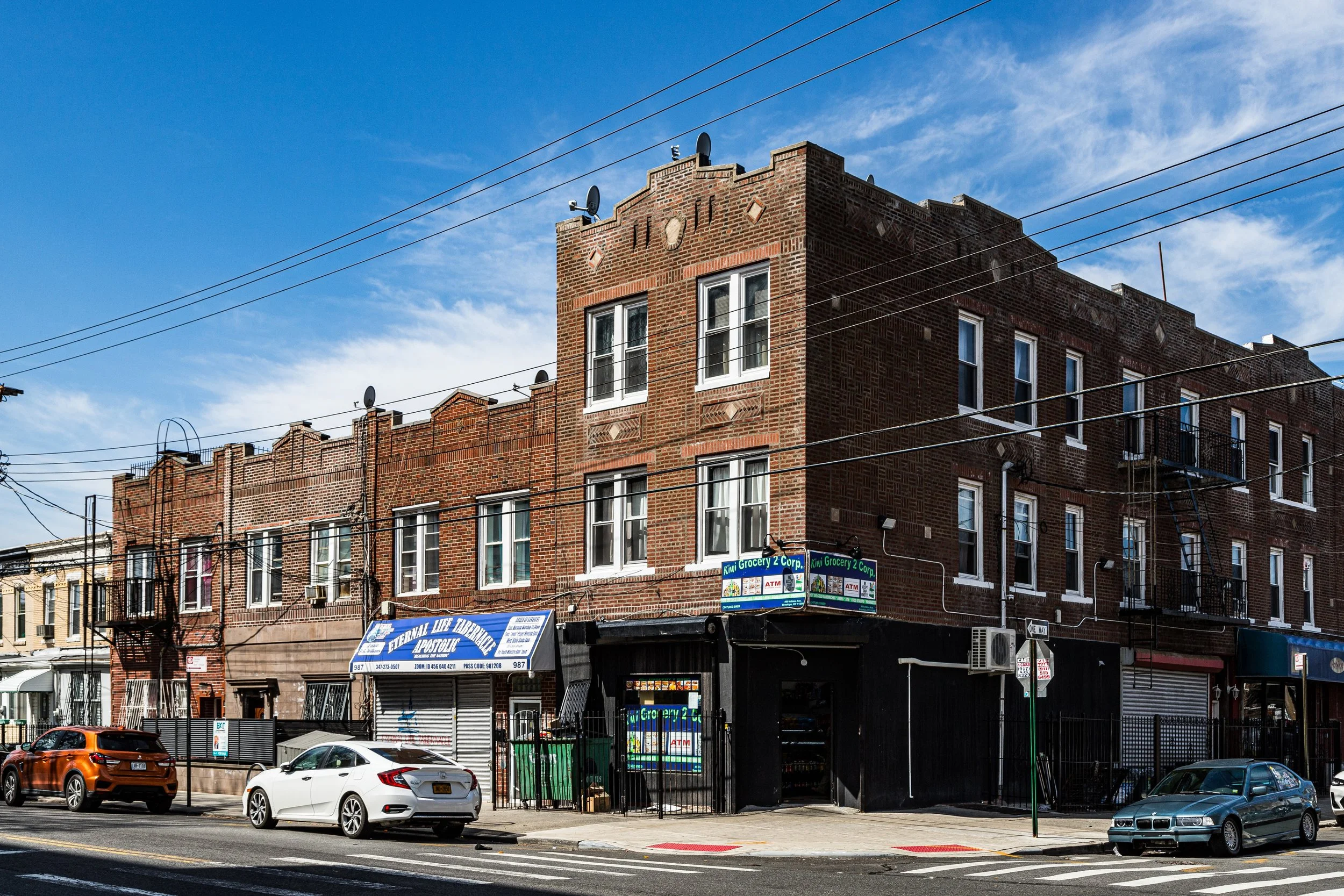 A multi-story brick building with a blue sky background and some white clouds, with parking cars in front. The building has several windows, a fire escape, and small satellite dishes on the roof.