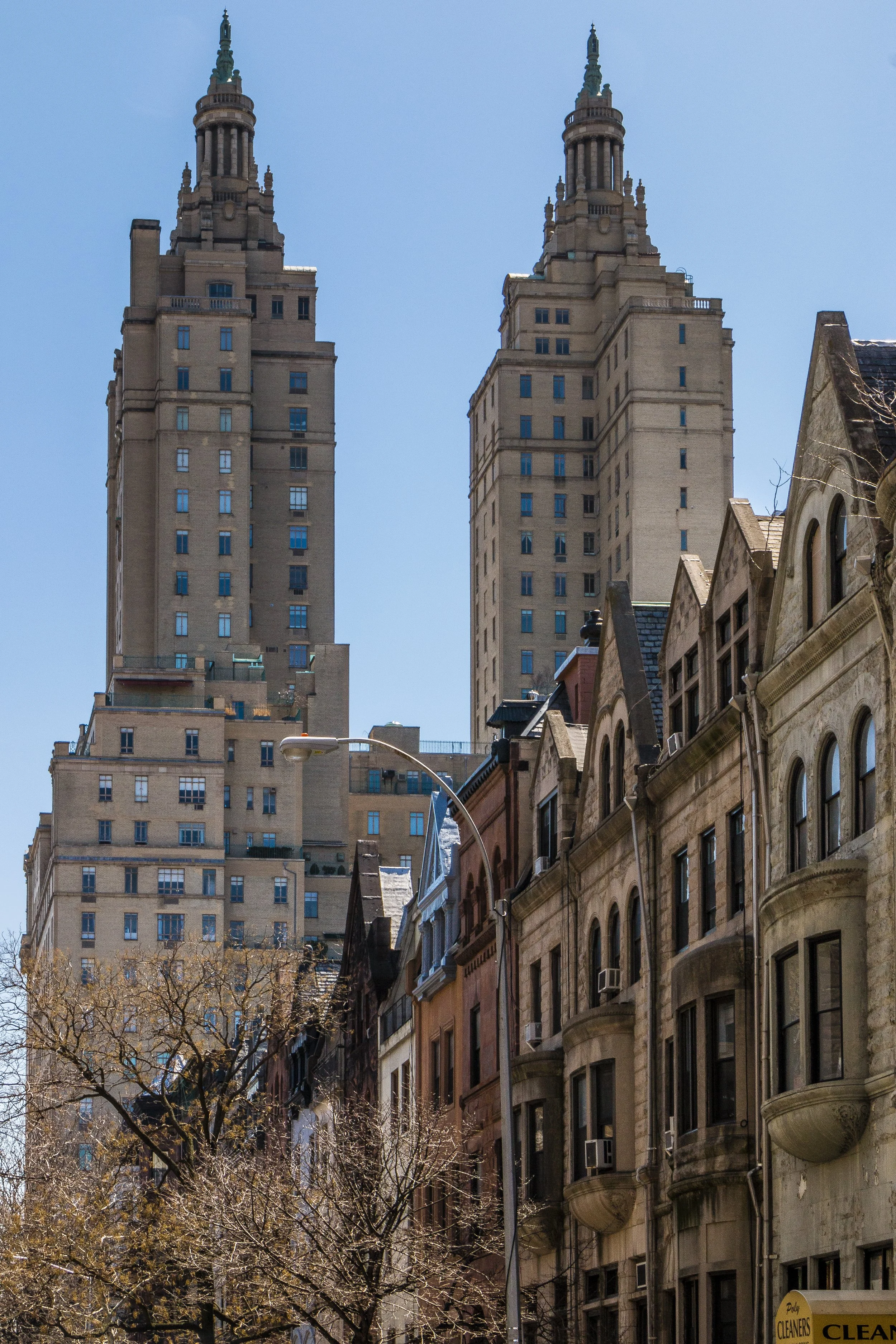 Two tall, historic skyscrapers with green spires rise above row houses in an urban street scene on a clear, sunny day.