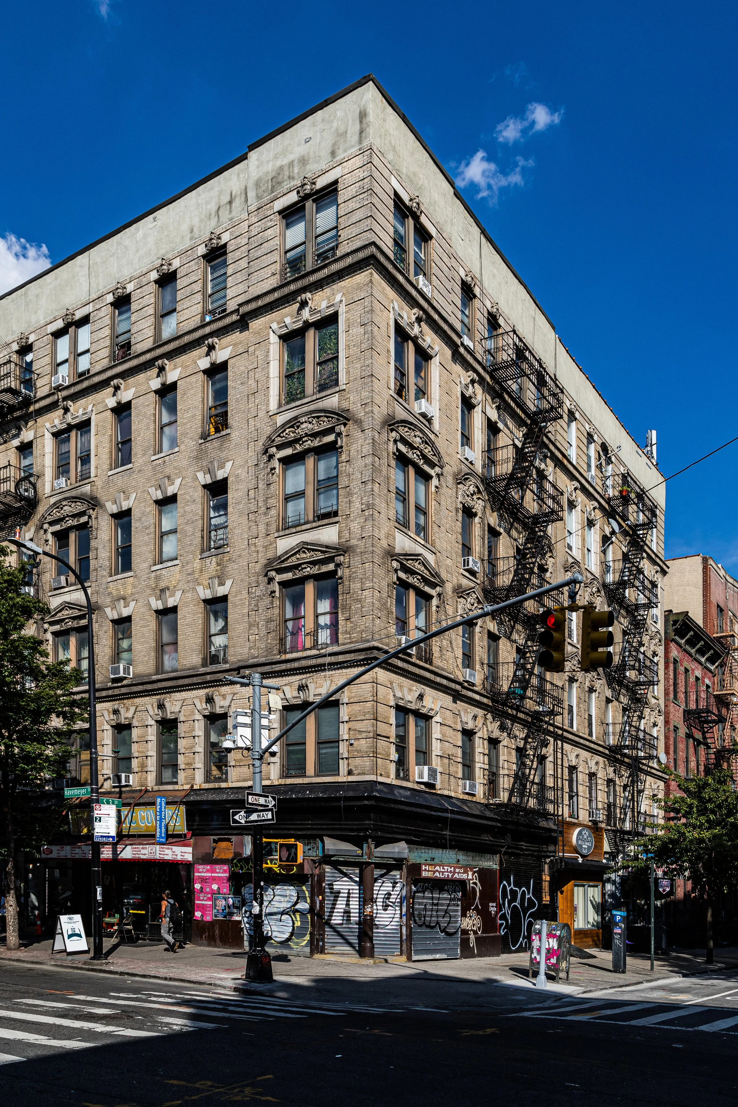 A multi-story brick building with fire escapes on the exterior, situated on a city street corner with traffic lights and pedestrians, under a blue sky with some clouds.